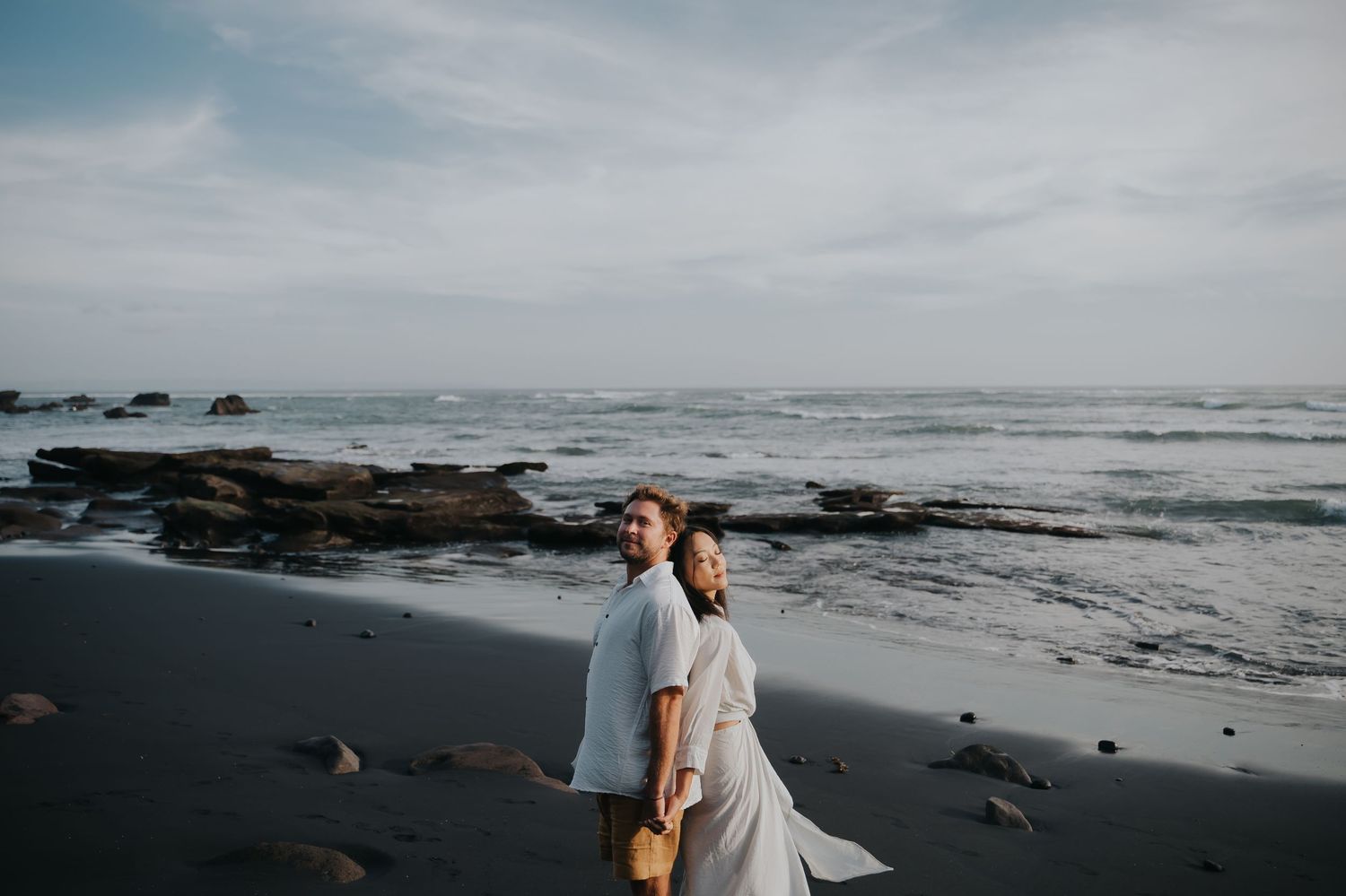 A couple walks along a misty black sand beach with rocky outcrops and ocean waves in the background.