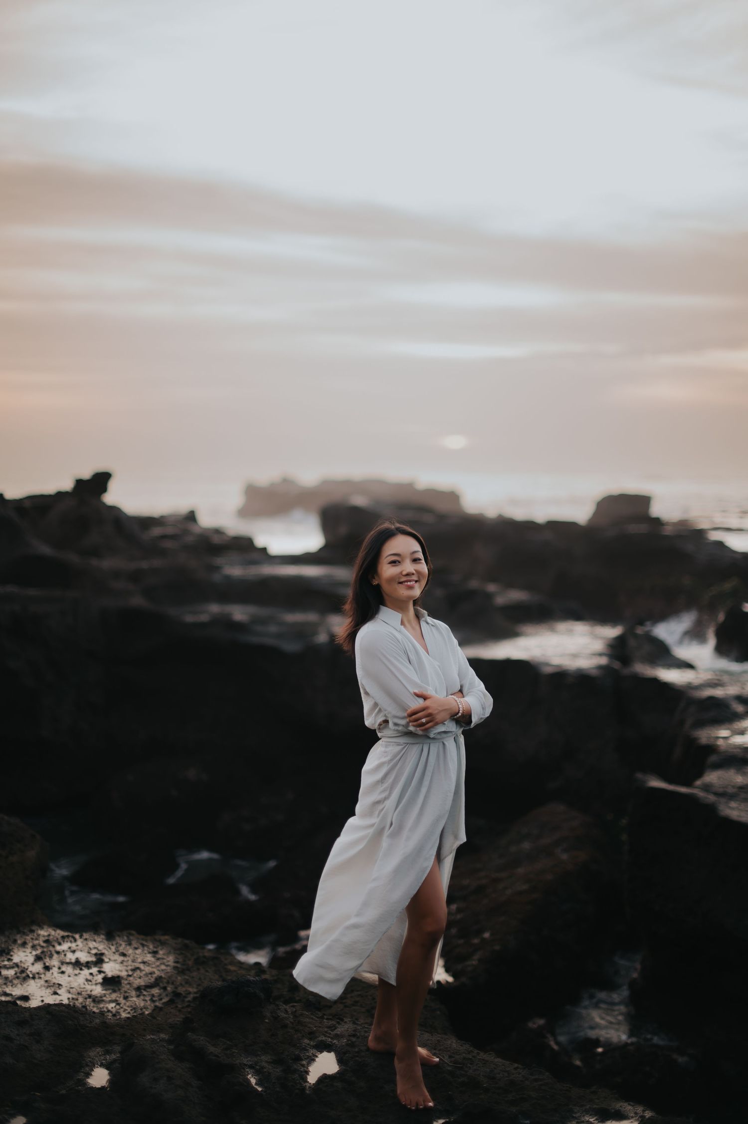 A person in a white dress stands on dark rocky coastal cliffs at sunset creating a dramatic silhouette against the sky.