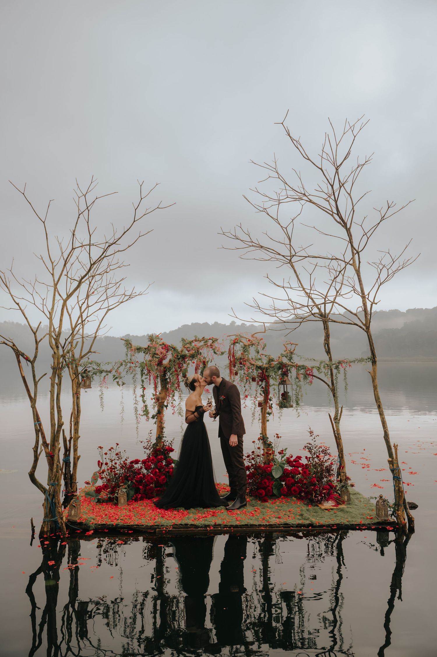 Moody wedding scene with bare trees and red floral arrangements rising from a misty lake.