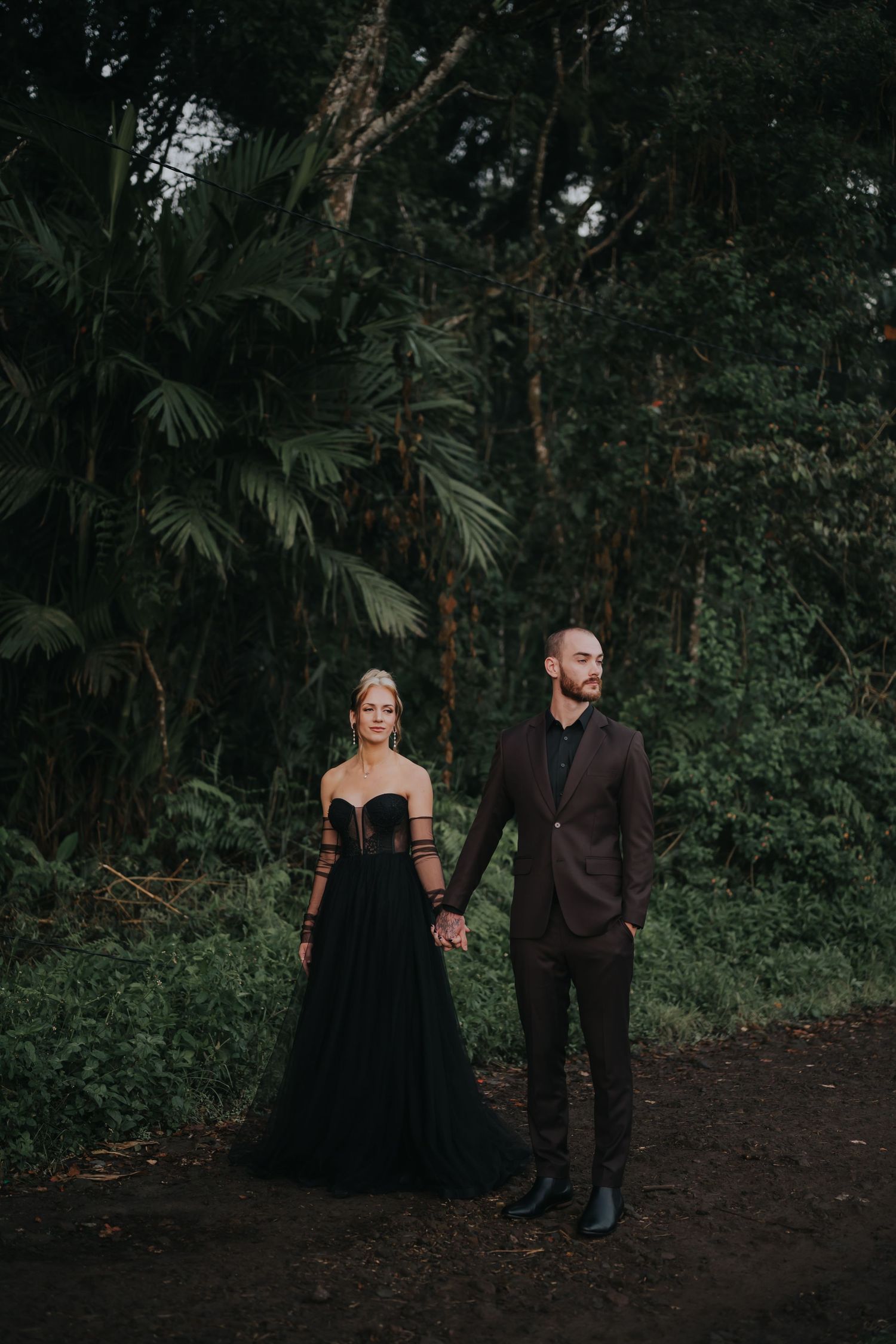 A couple in formal black attire poses together on a forest path surrounded by lush tropical foliage.
