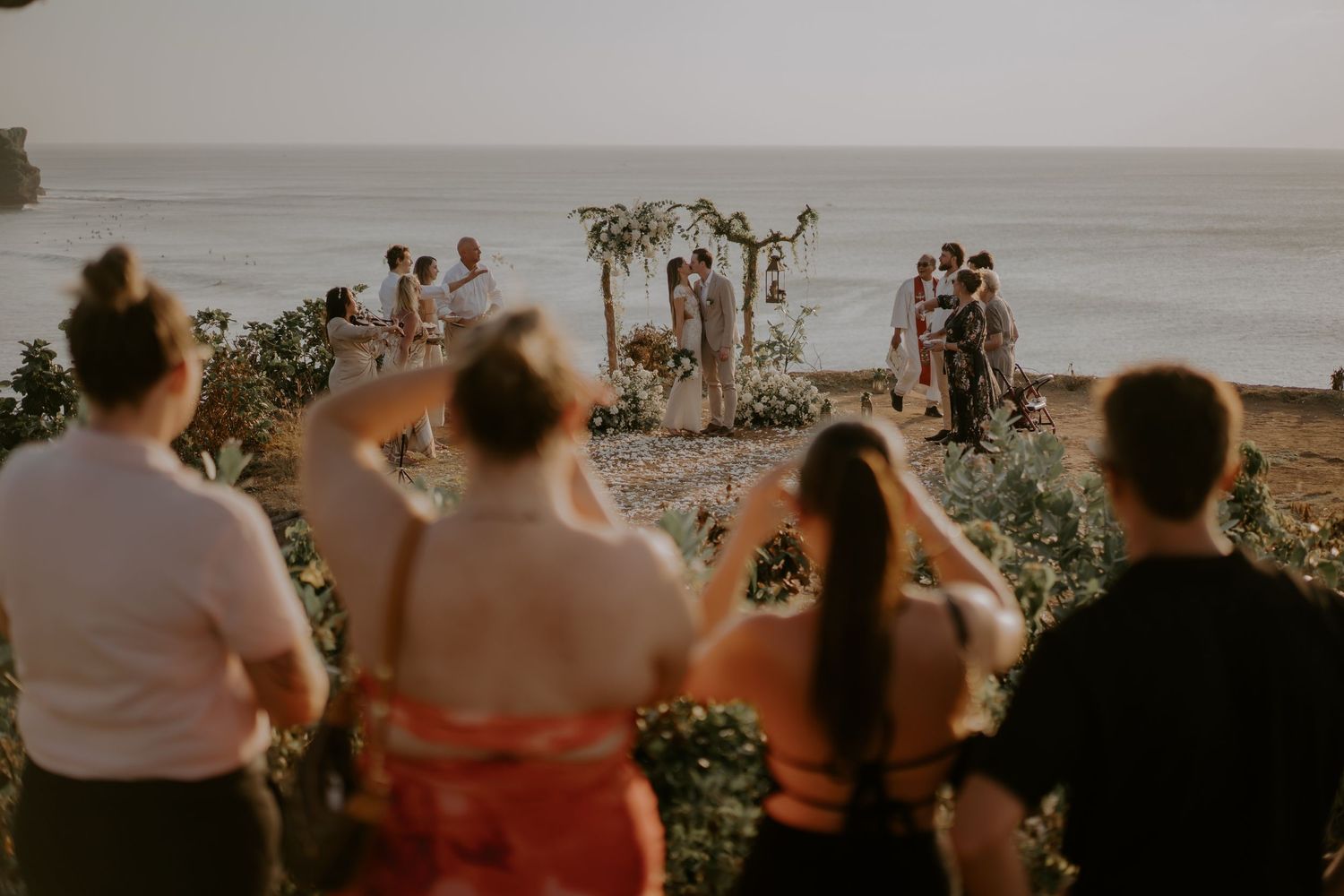 Wedding guests watch a sunset beach ceremony from a cliff overlooking the ocean with a floral arch setup.