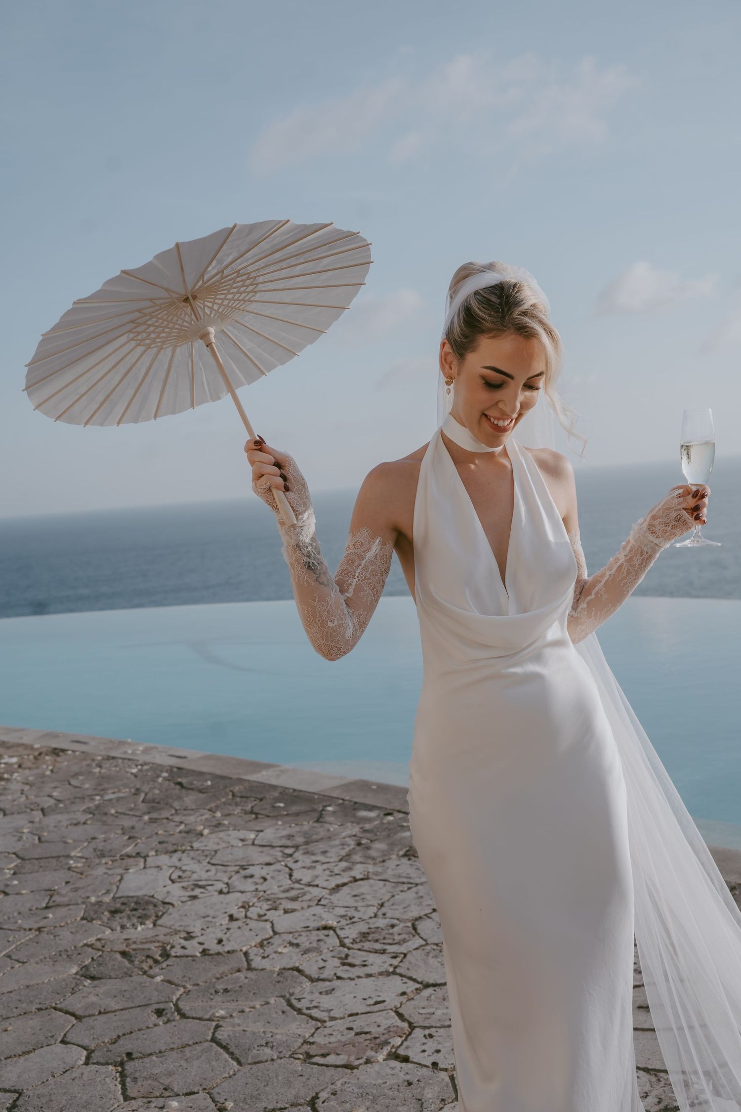 Elegant bride in white gown poses with parasol and champagne against coastal scenery.