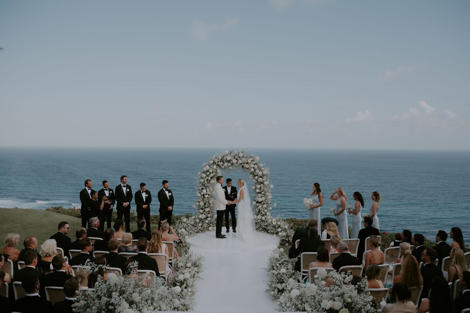 Oceanfront wedding ceremony under a floral arch with guests seated on either side.