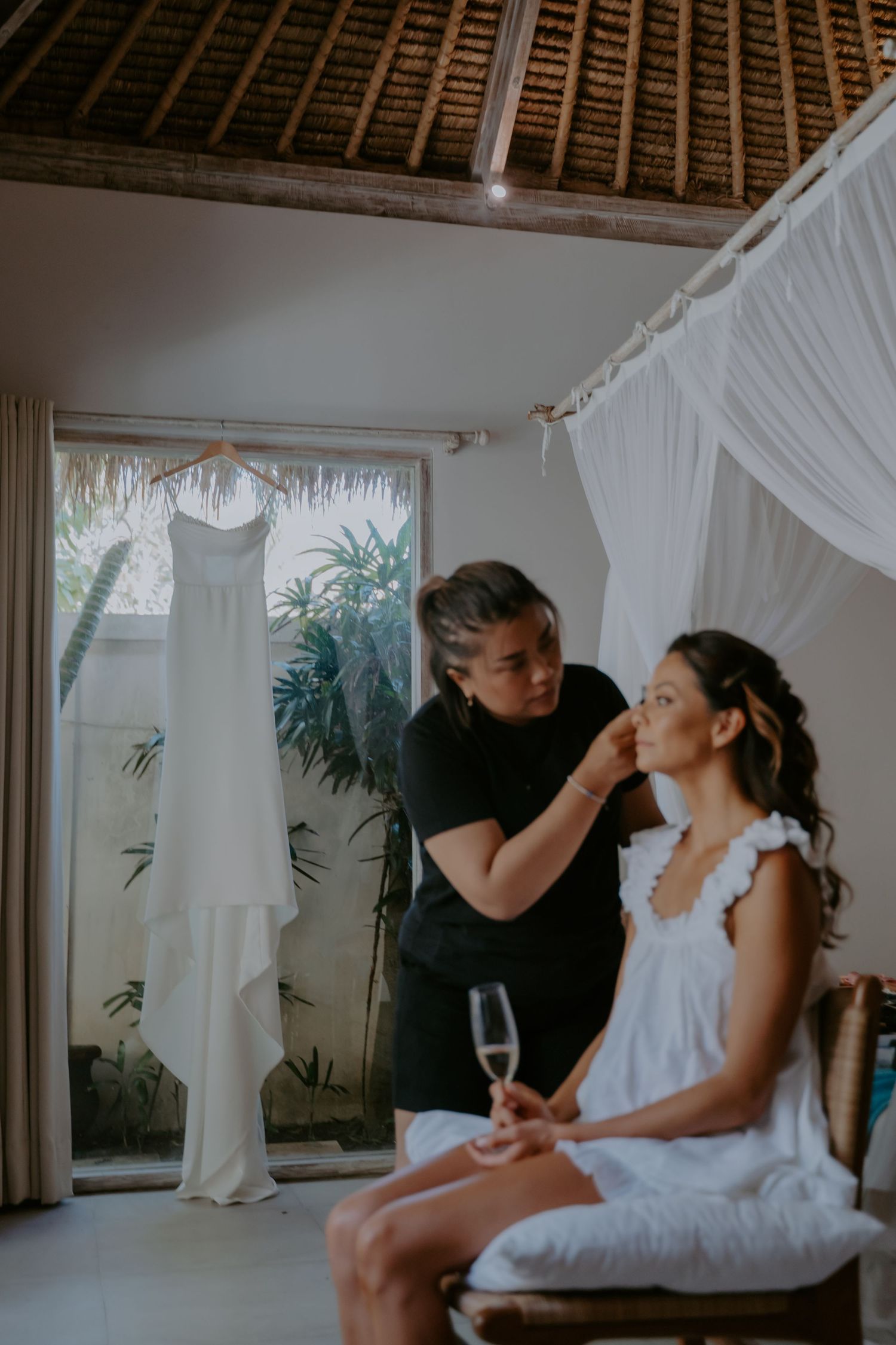 Romantic wedding preparation scene with white curtains and tropical plants visible through windows.
