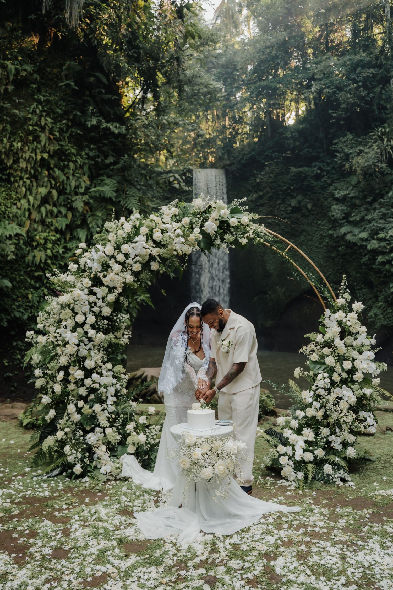 Bride and groom cutting wedding cake under a white floral arch with a waterfall backdrop in Bali.
