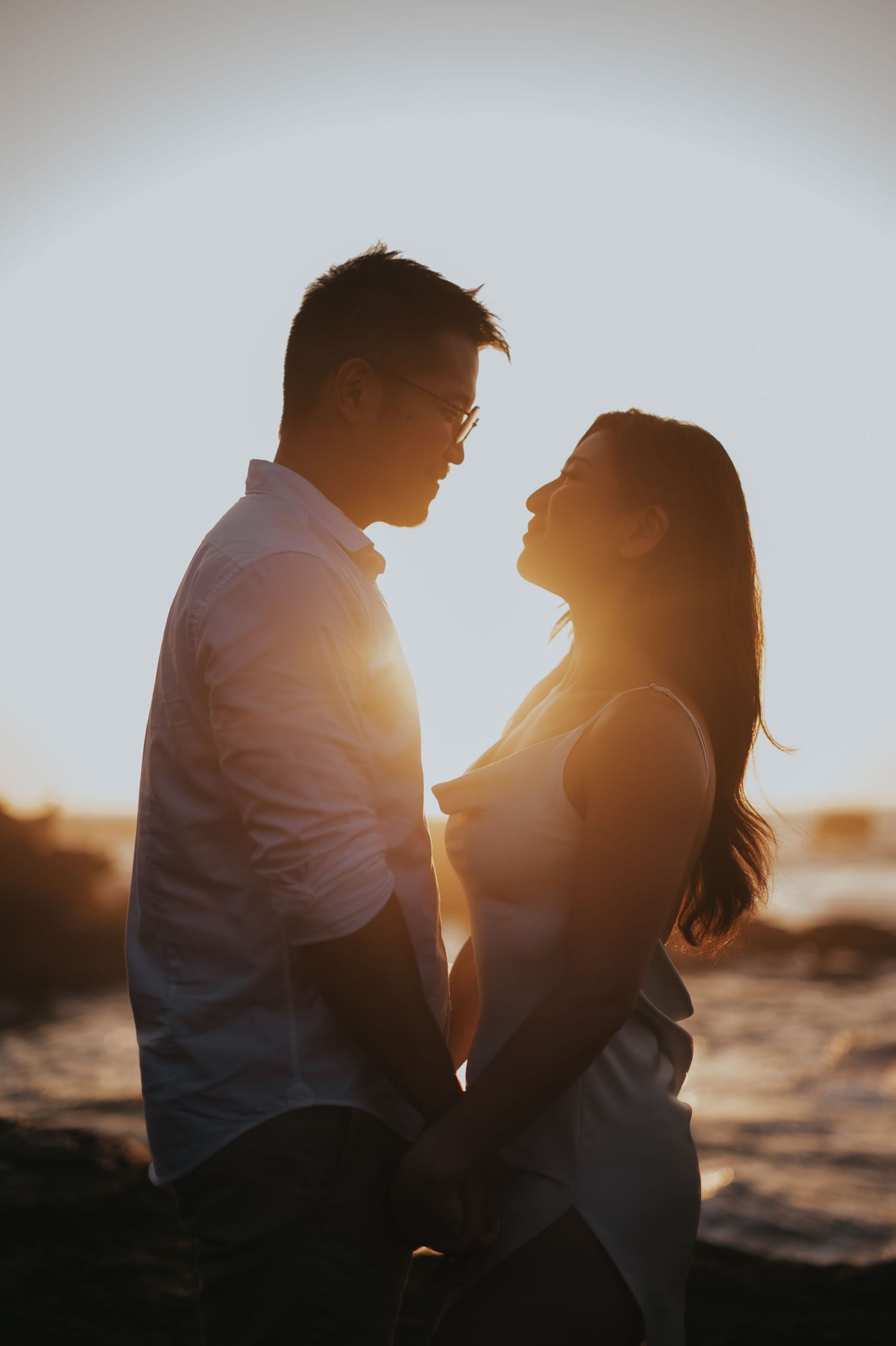 Romantic couple silhouetted against golden sunset light by the ocean, sharing an intimate moment while holding hands.
