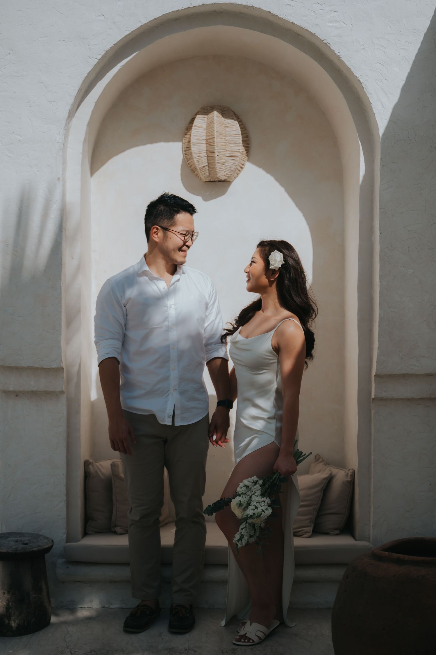 Couple in white attire standing under an arched alcove, smiling at each other in a romantic Mediterranean setting.