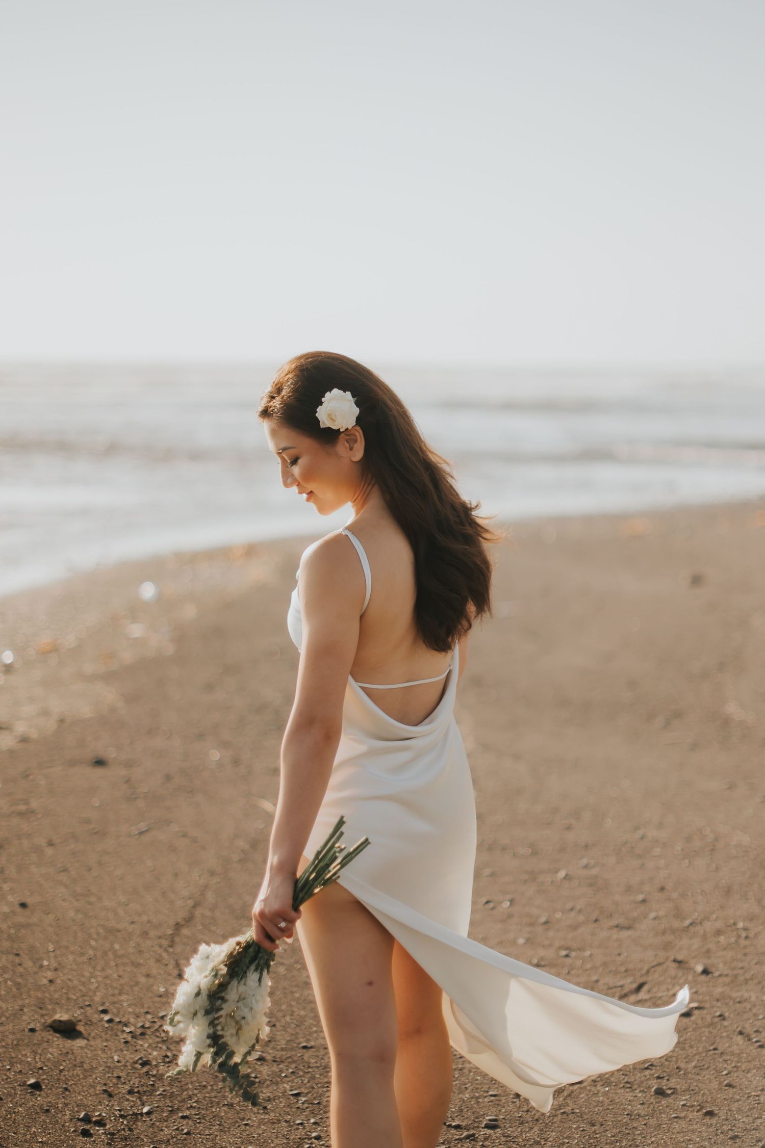 Woman in white backless dress holding bouquet walks on sandy beach with ocean waves, white flower in dark hair.
