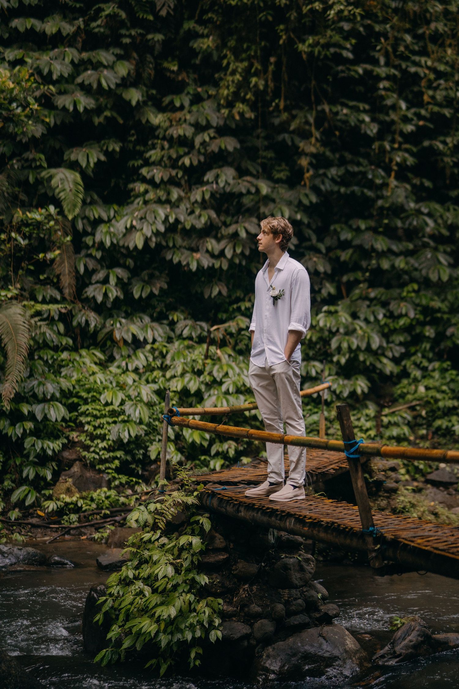 Someone in casual white attire stands on a wooden footbridge over a stream surrounded by lush green foliage.