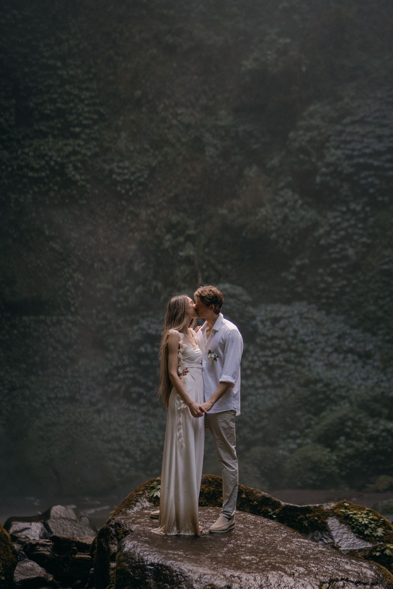 A couple shares a romantic moment on a rocky cliff edge overlooking a misty mountain landscape at dusk.