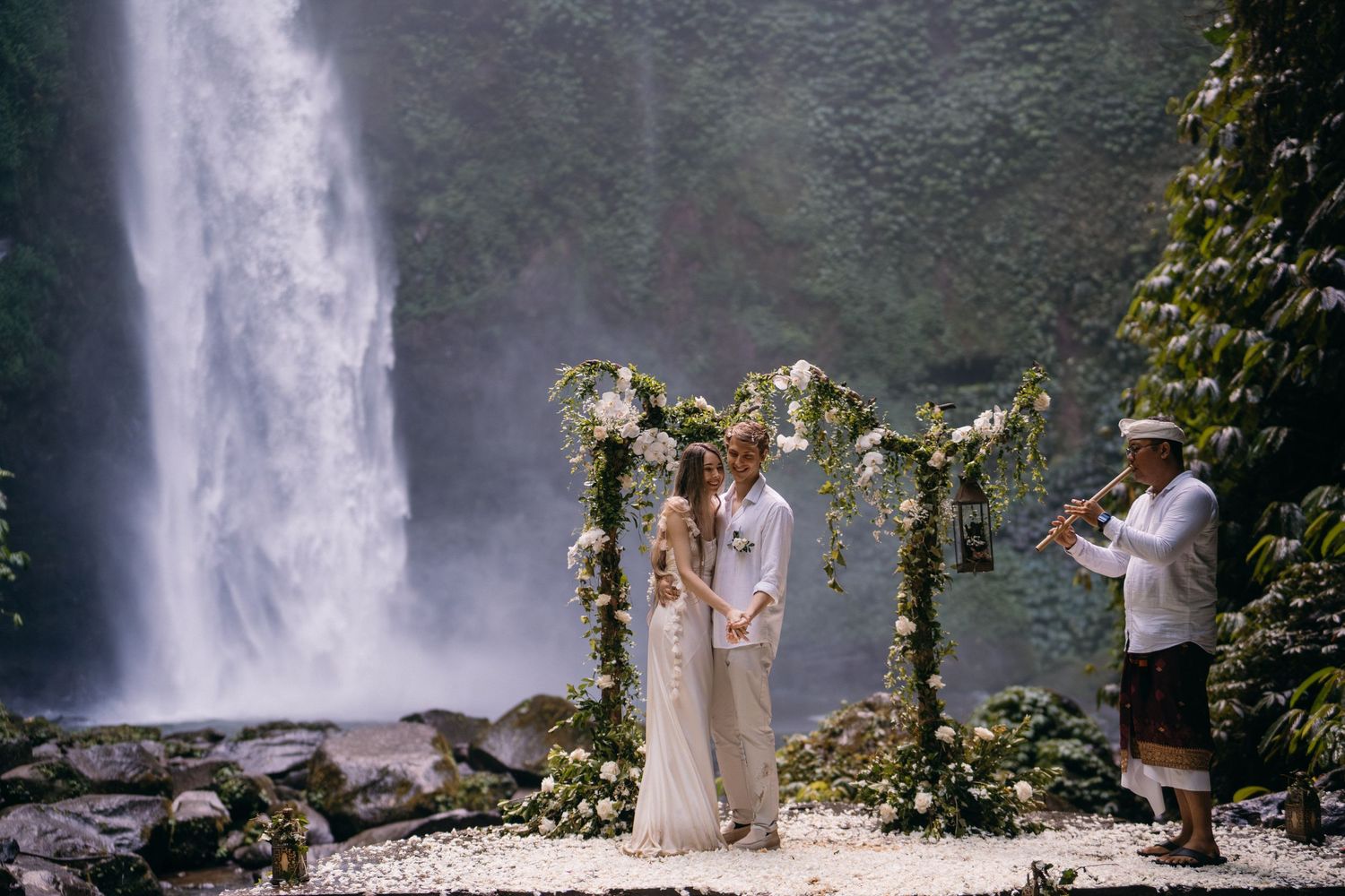 Wedding ceremony taking place in front of a majestic waterfall with lush greenery surrounding the scene.