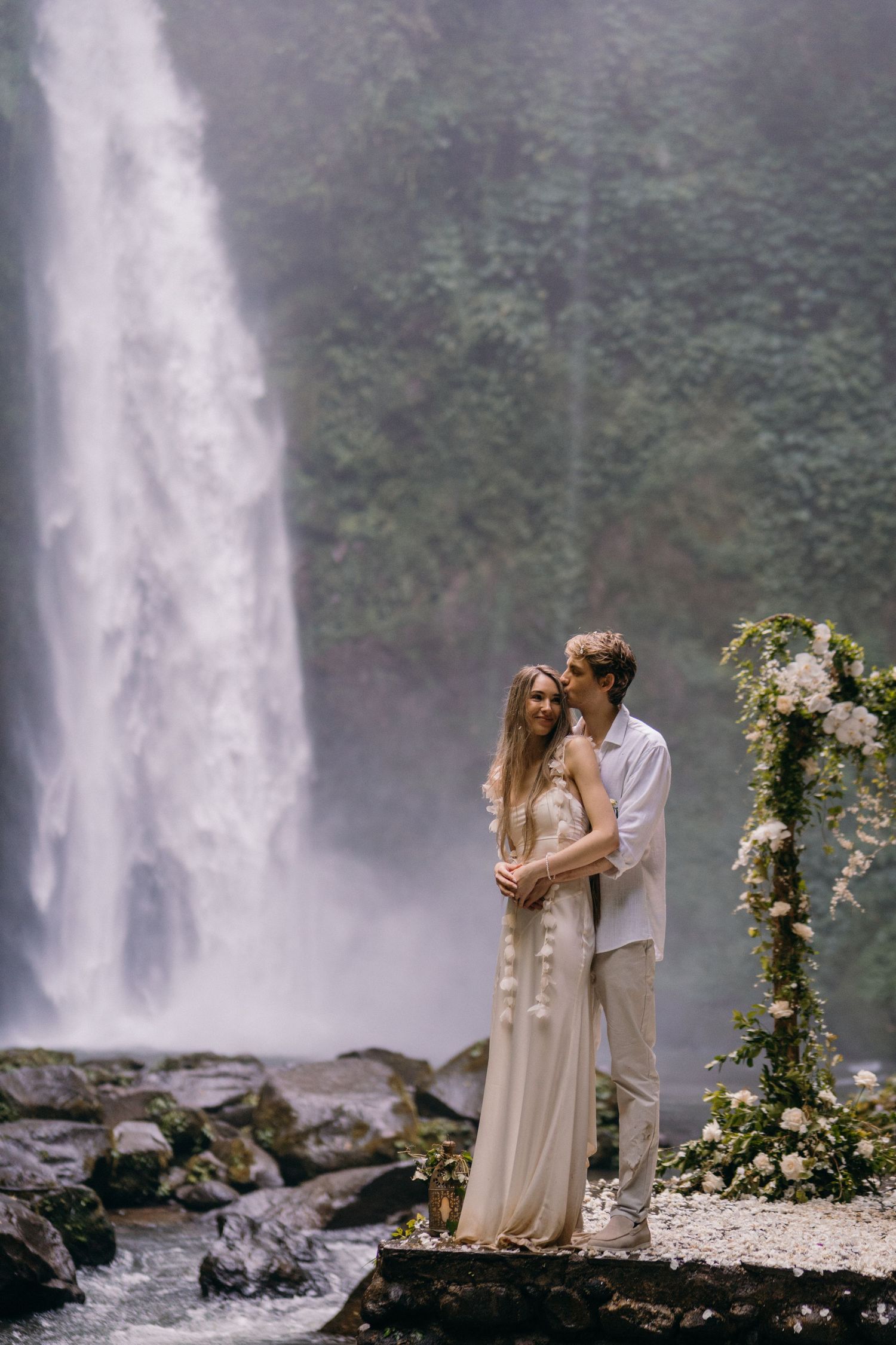 A romantic couple embraces near a majestic waterfall with a floral arch and misty atmosphere in the background.