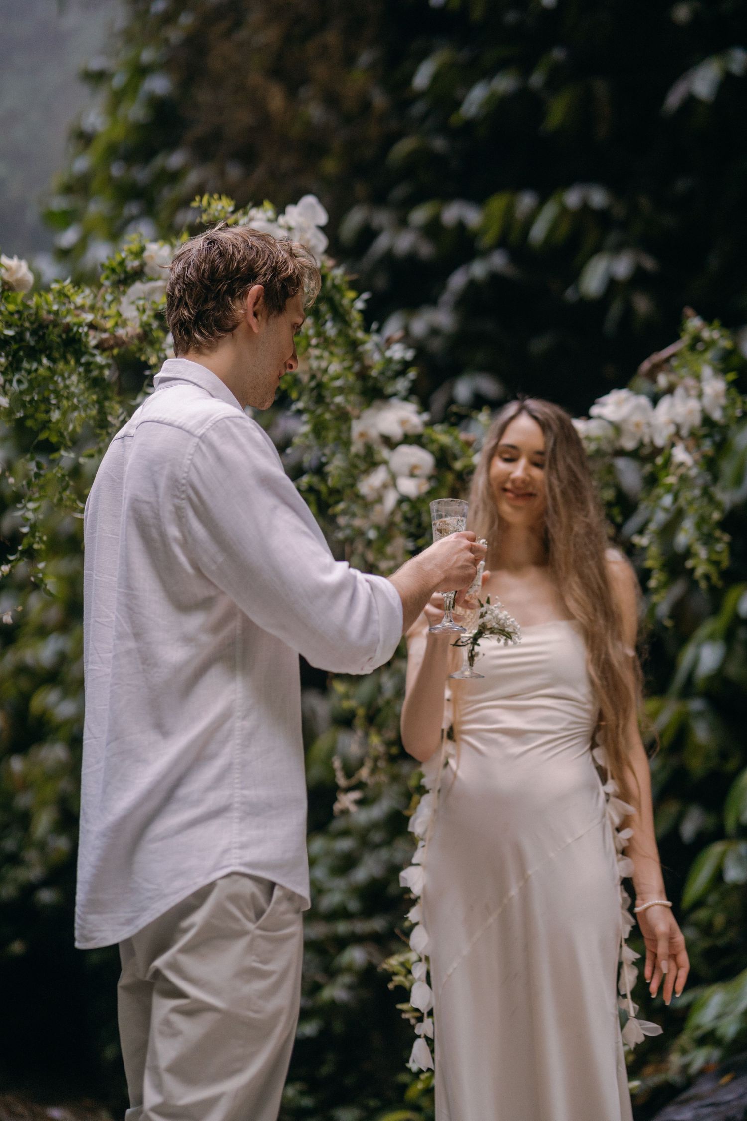 A couple in light-colored attire dance together in a garden setting with flowering vines in the background.
