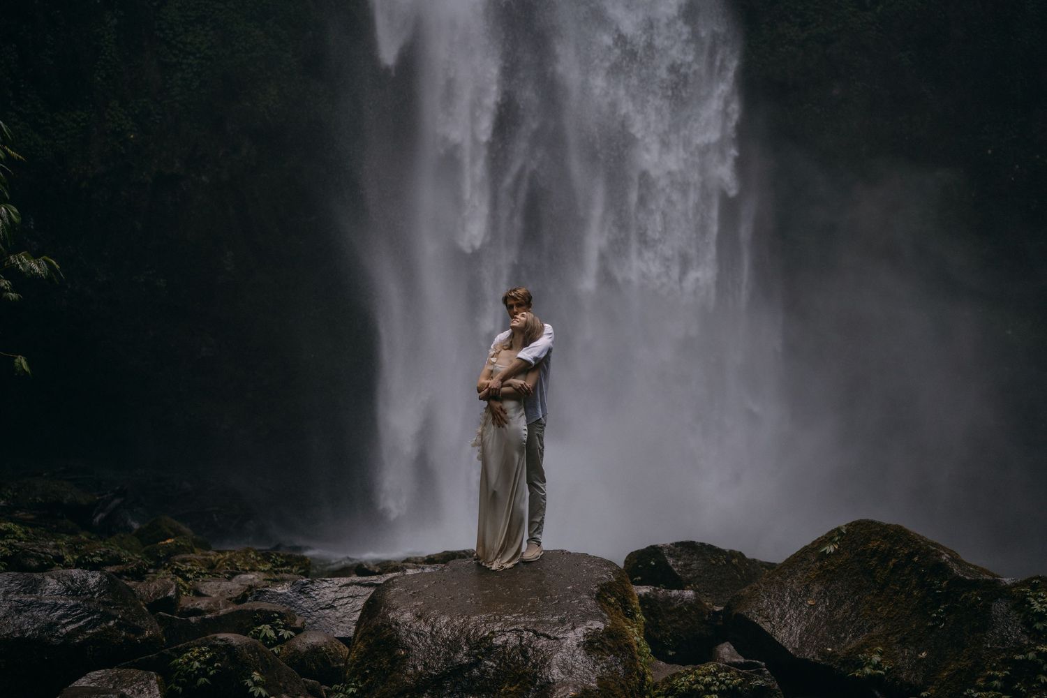 A misty waterfall cascades down dark rocky cliffs as a silhouetted figure stands near its powerful flow.