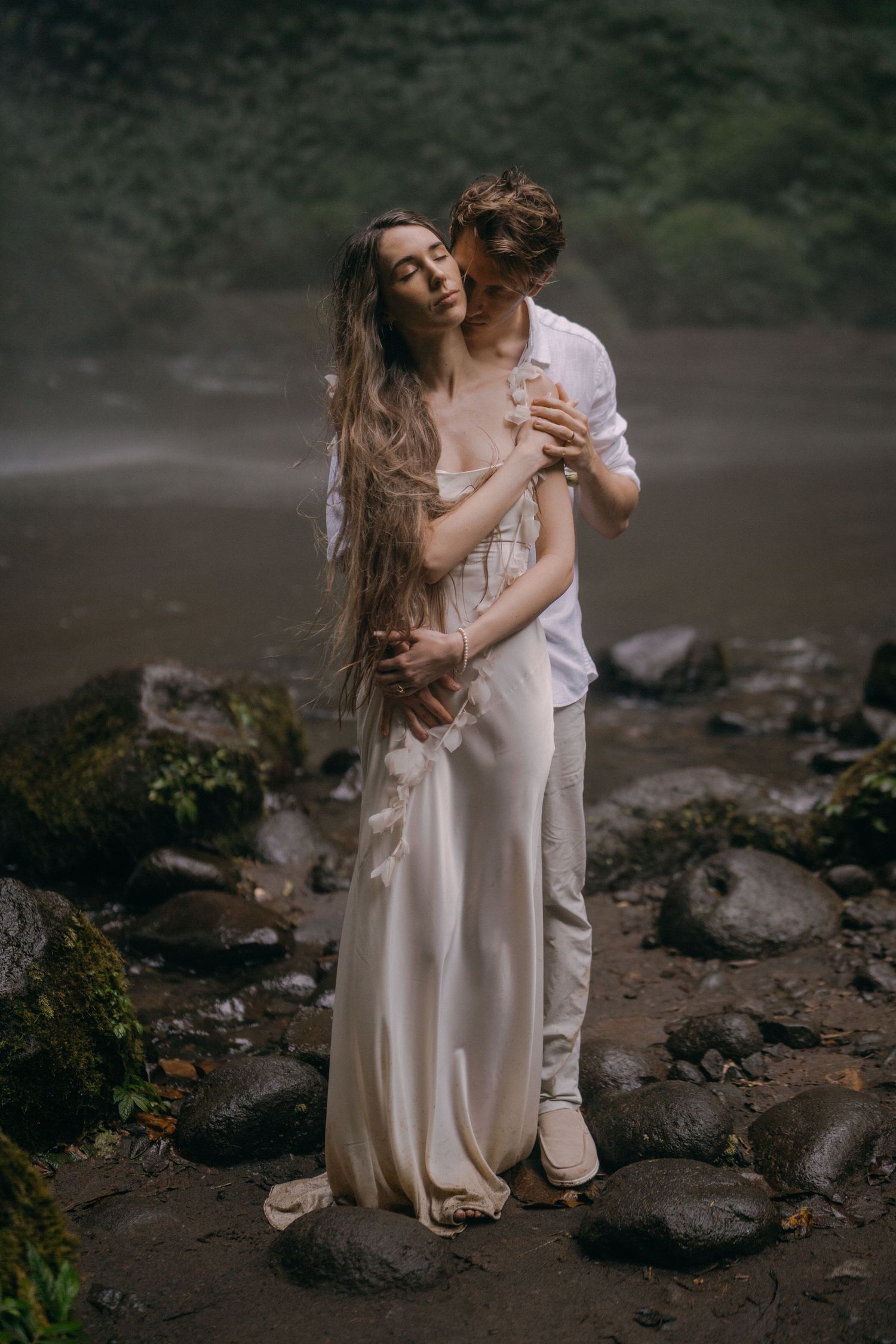 A couple in flowing white outfits embraces tenderly on a rocky beach landscape at dusk.
