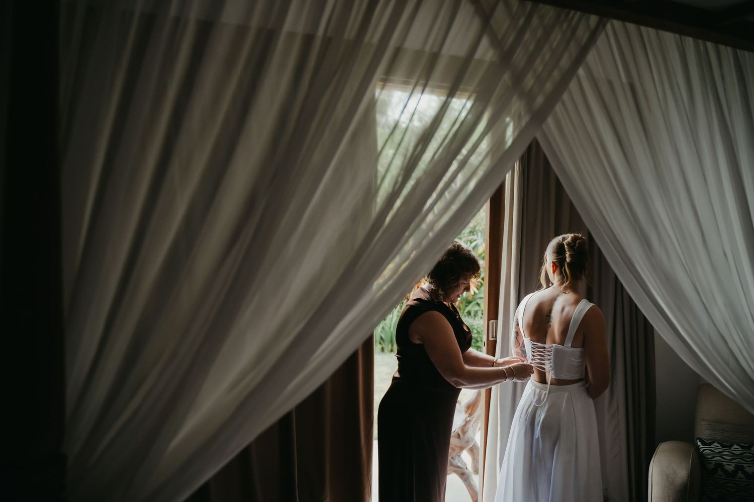 Helper laces up brides corset wedding dress near sheer curtains in soft natural light.