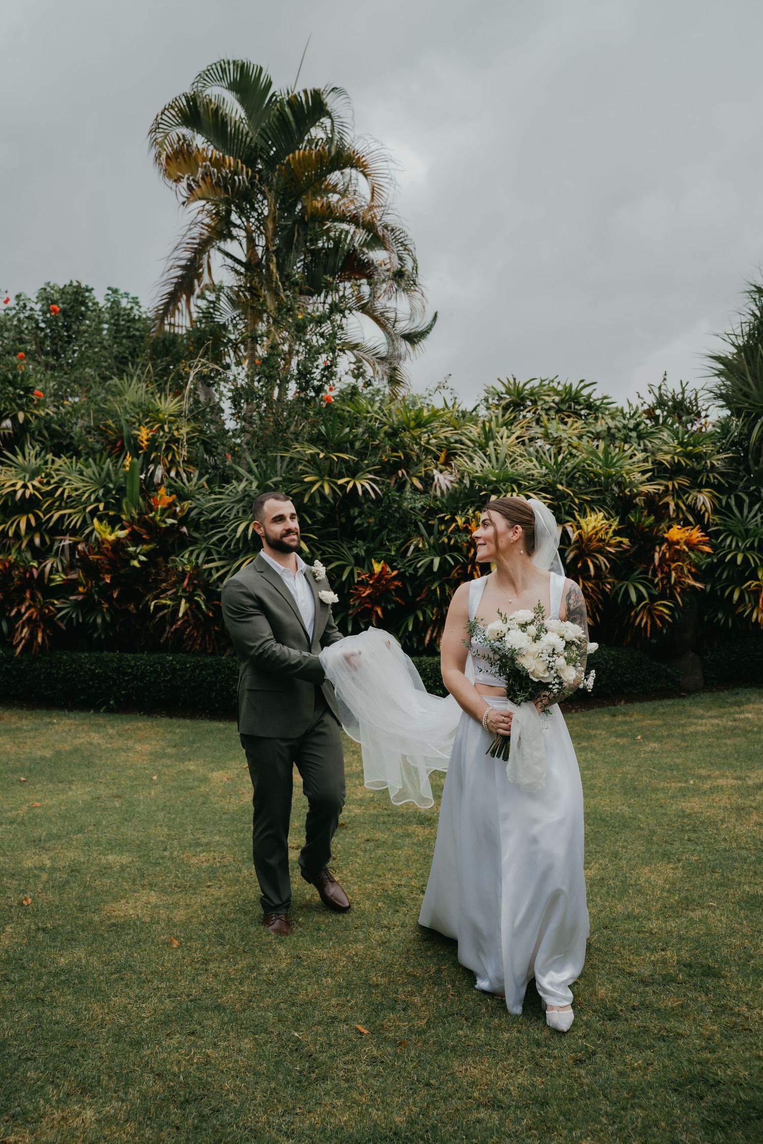 Groom playfully holding brides veil as she laughs in tropical garden wedding portrait outdoors.