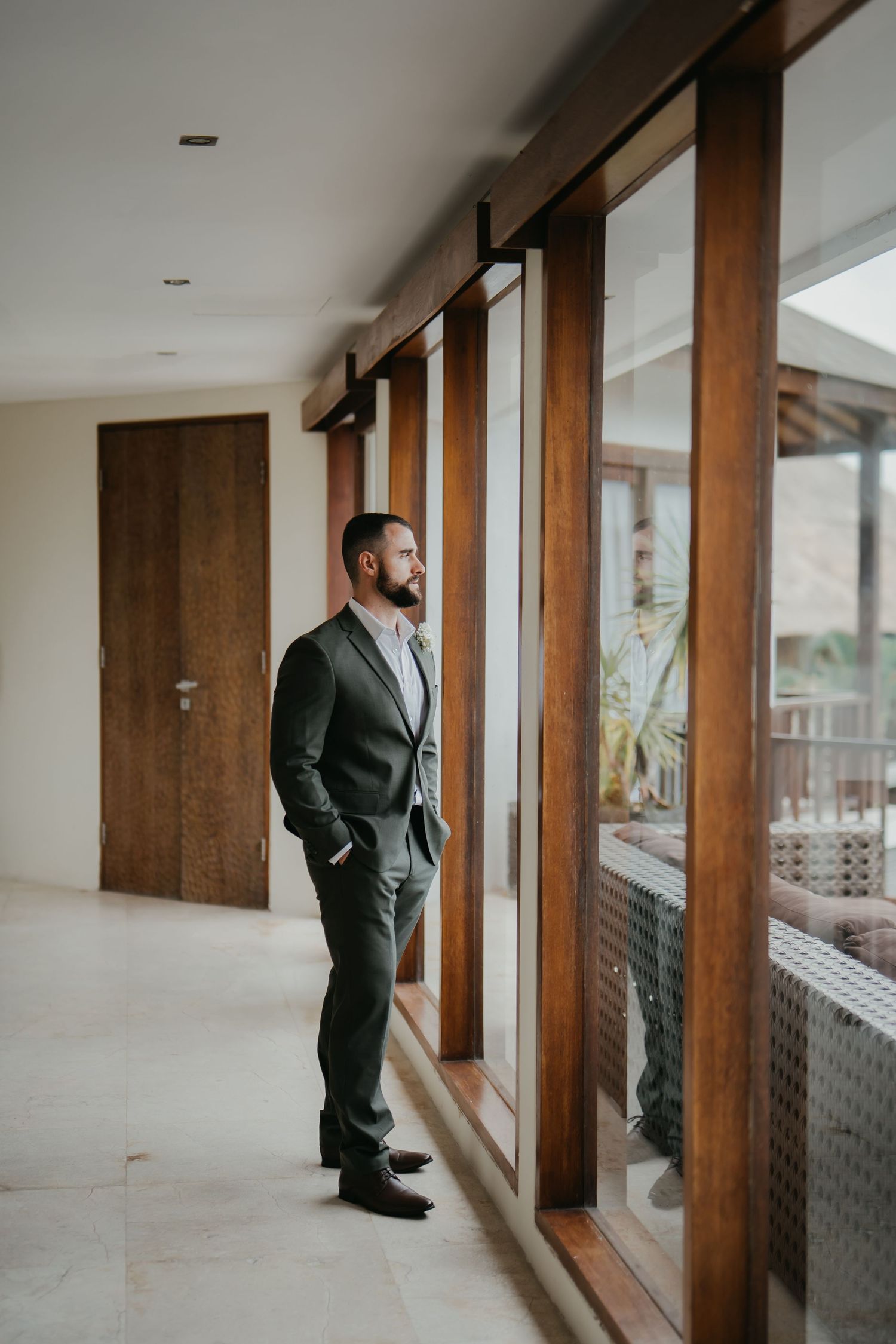 Stylish groom in charcoal suit stands contemplatively by large wooden framed glass windows in a modern villa.
