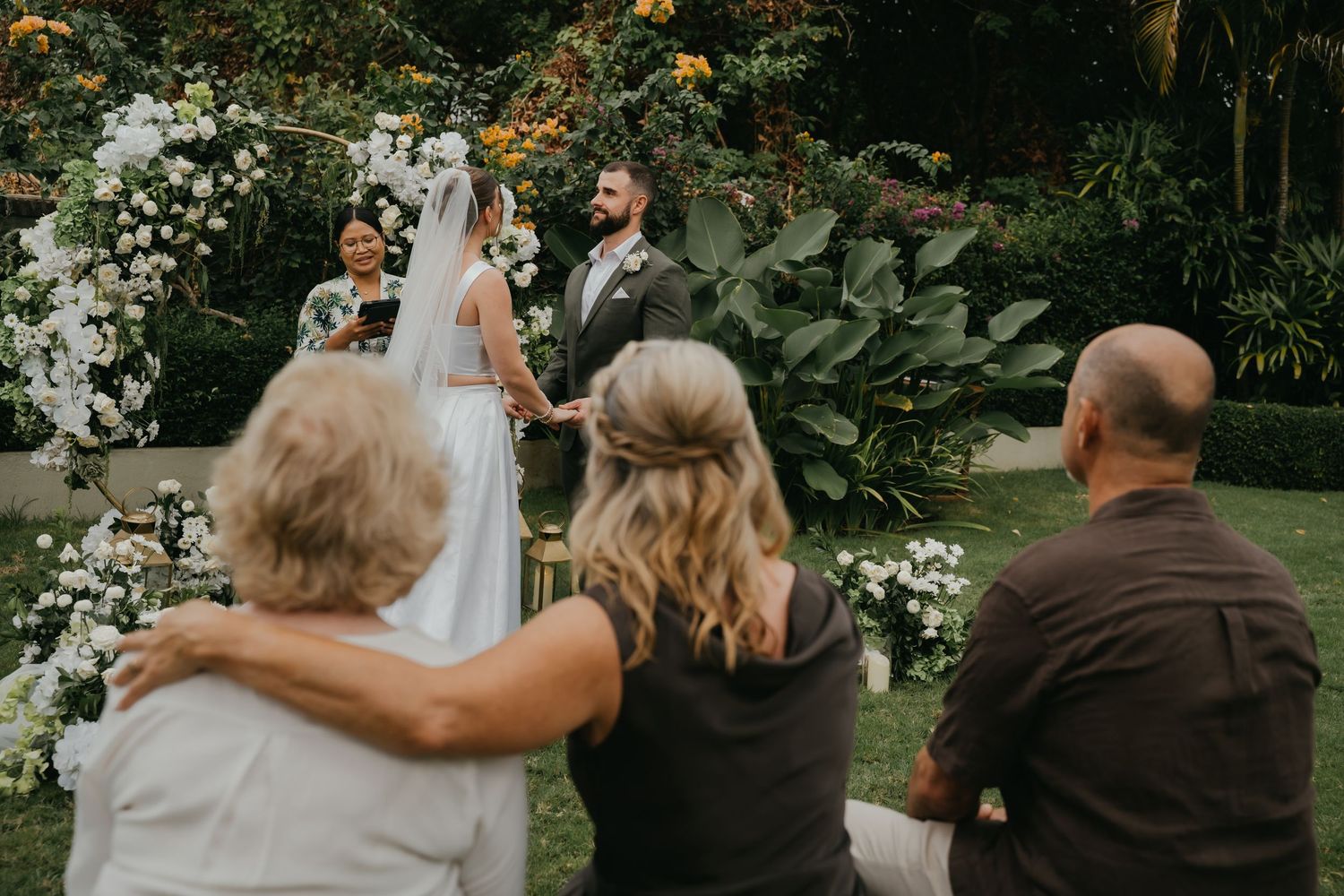 Wedding guests embrace while watching bride and groom exchange vows under white floral arch in garden.