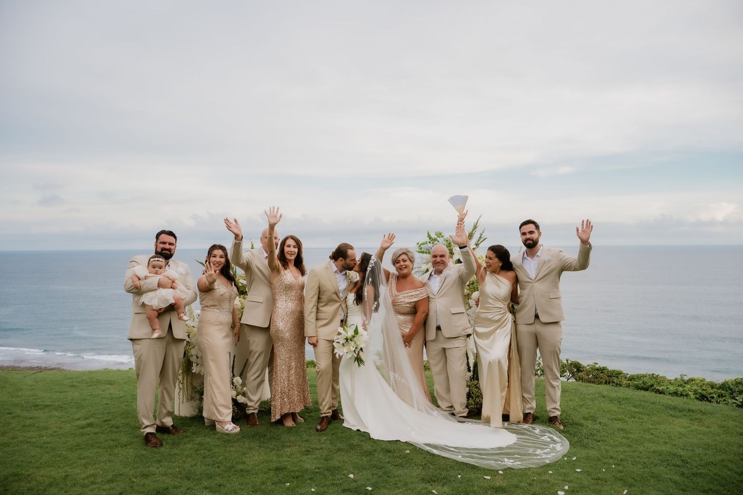 Wedding party in neutral tones cheers together on cliffside lawn with ocean in background.