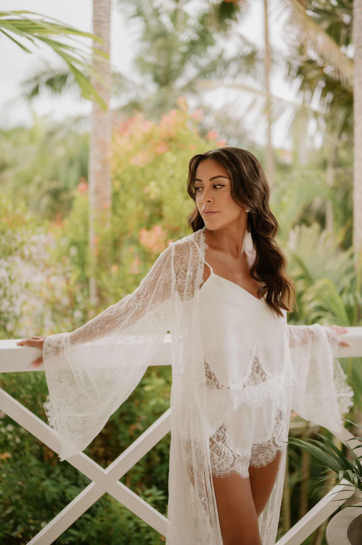 Bride in white lace robe standing on tropical balcony with palm trees in background.