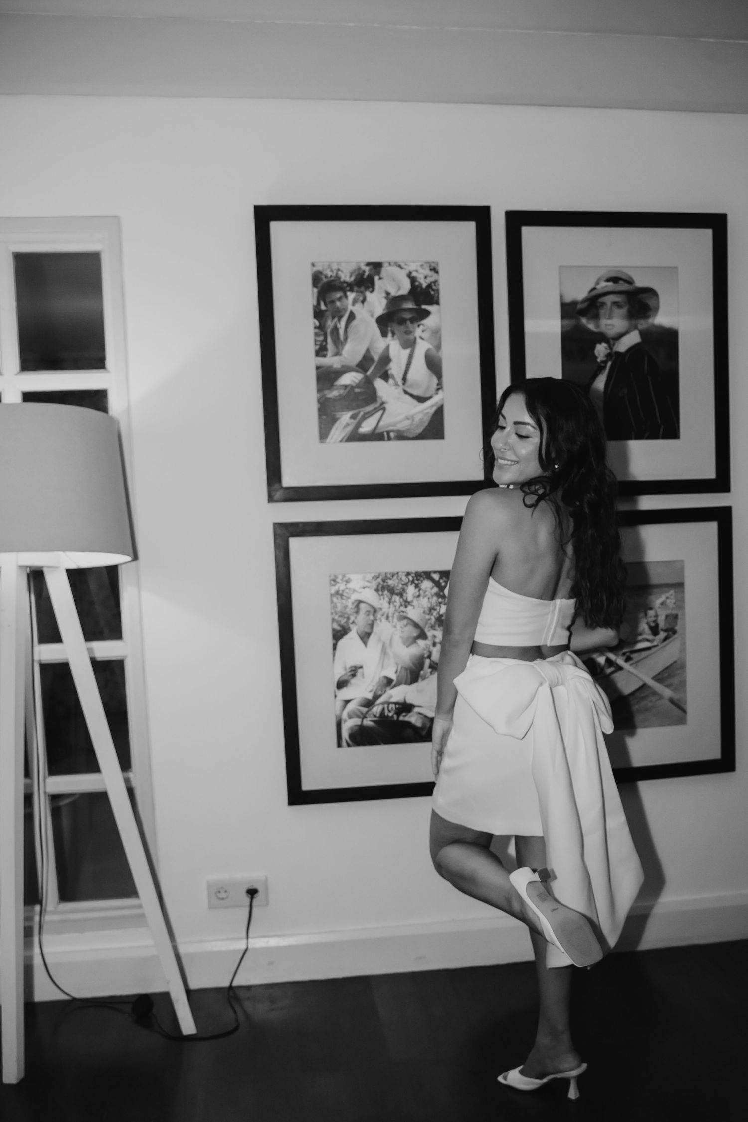 Black and white photo of woman in white crop top and skirt posing by framed wall art gallery in stylish interior.