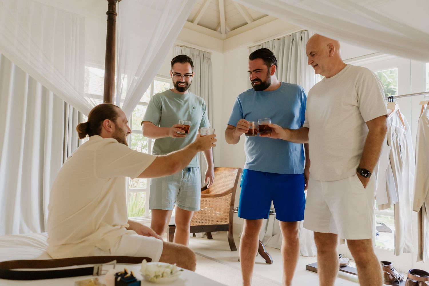 Four men in casual summer attire sharing drinks and conversation in an elegant white canopied room.