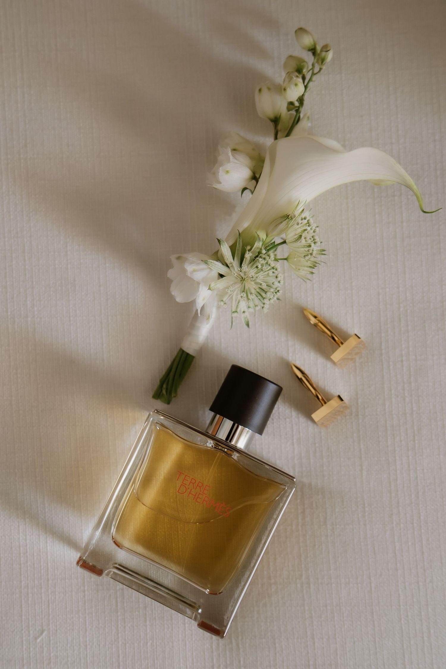 Elegant wedding flat lay with gold perfume bottle, white flowers, and gold cufflinks on a neutral background.