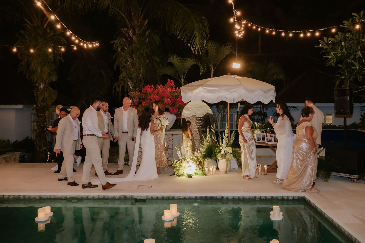 Wedding guests mingle poolside at night under string lights with floating candles on the water.