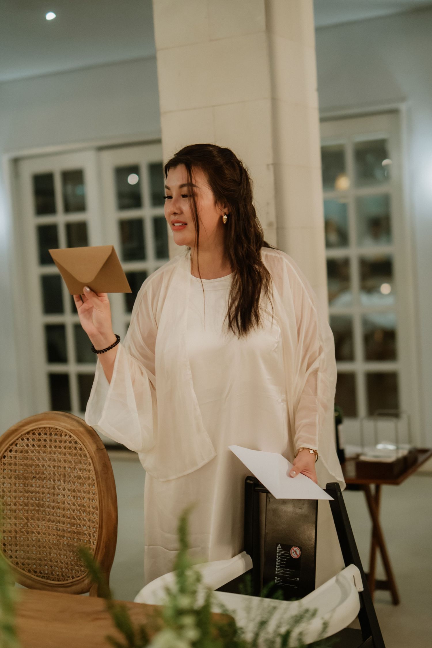 Elegant woman in cream attire holds an envelope while speaking at an indoor wedding reception event.