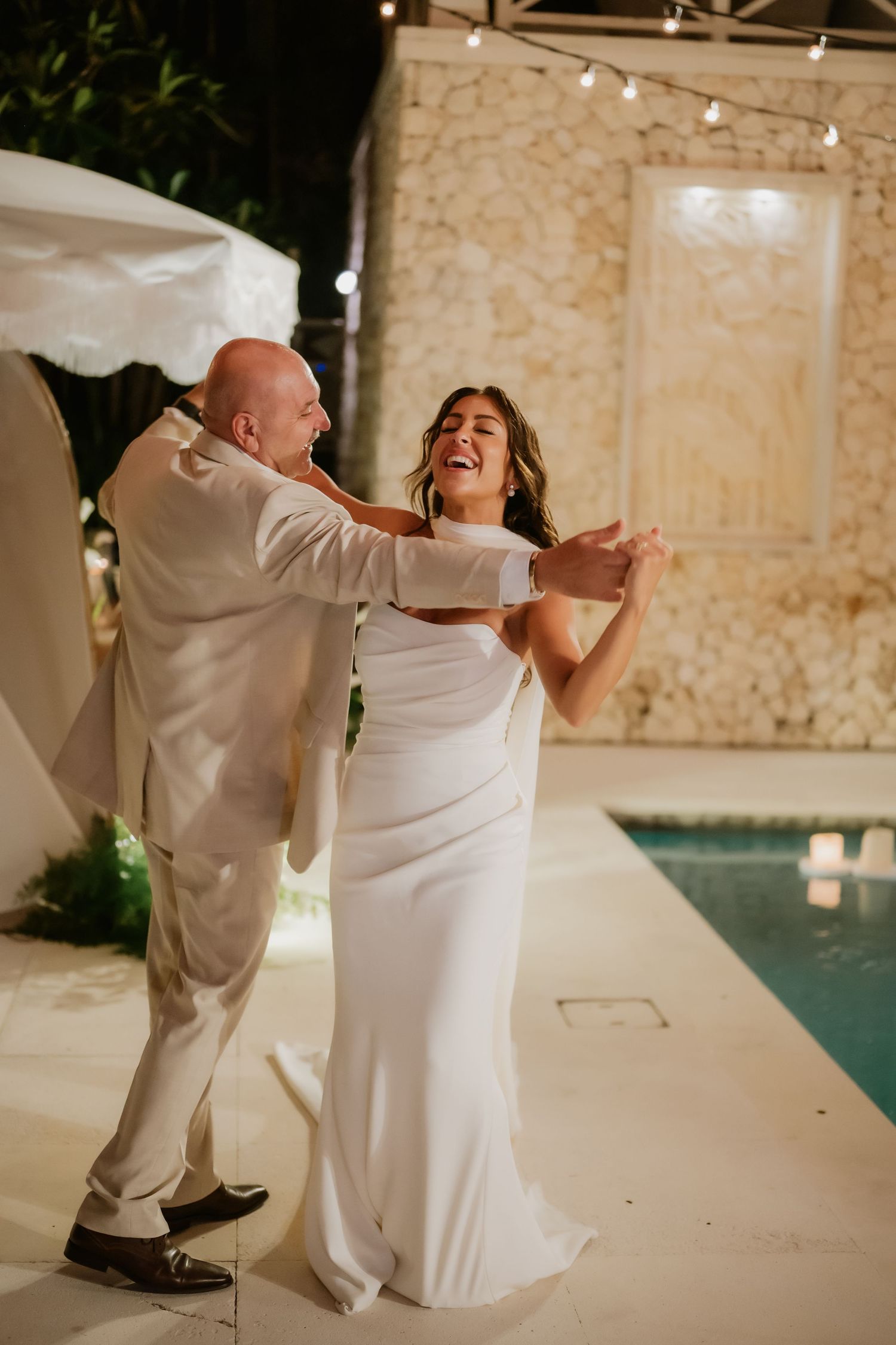 Joyful bride in white gown laughing with groom during first dance beside illuminated pool at night.