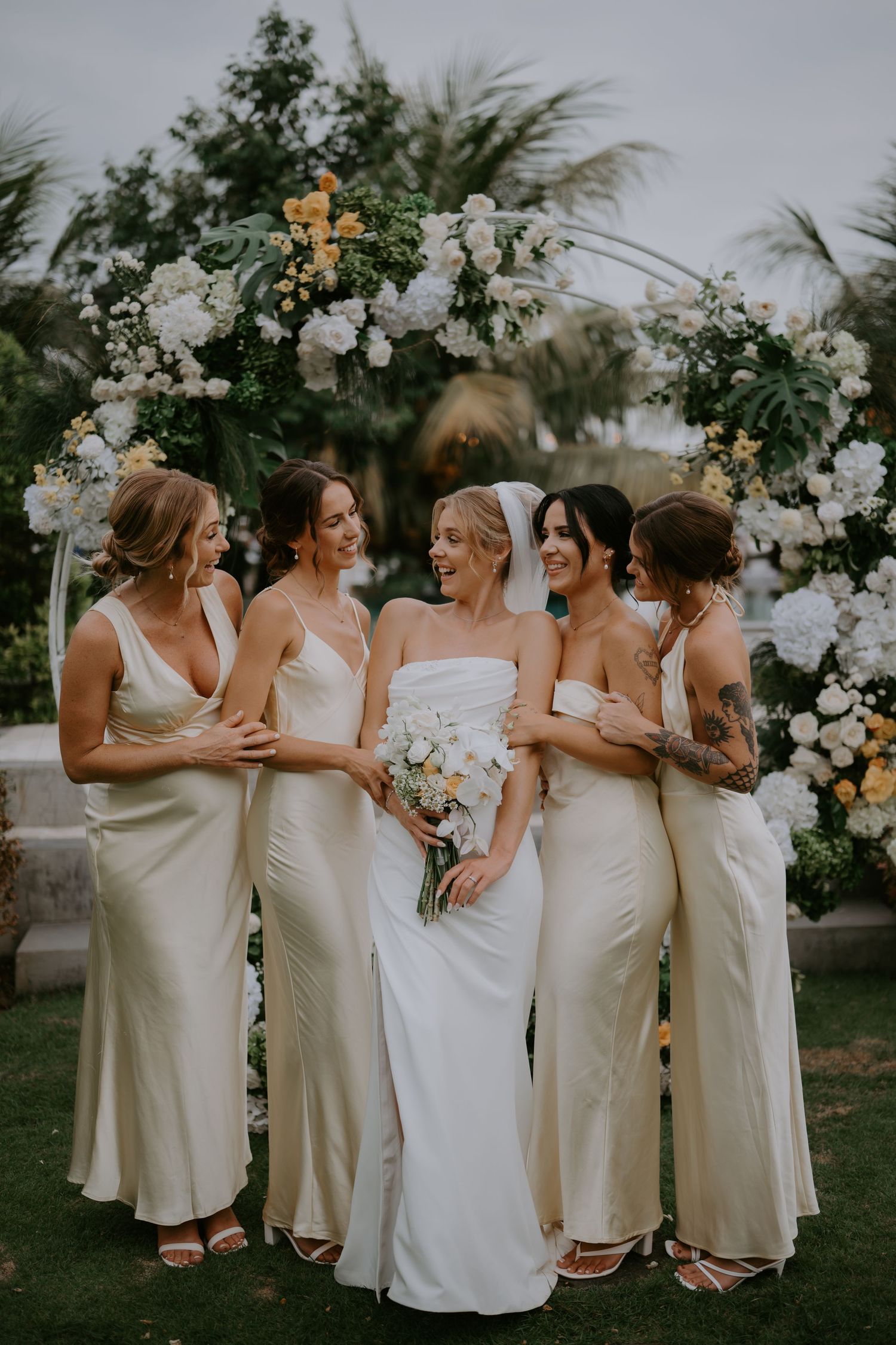 Bride with bouquet poses with four bridesmaids in champagne gowns under white floral arch.