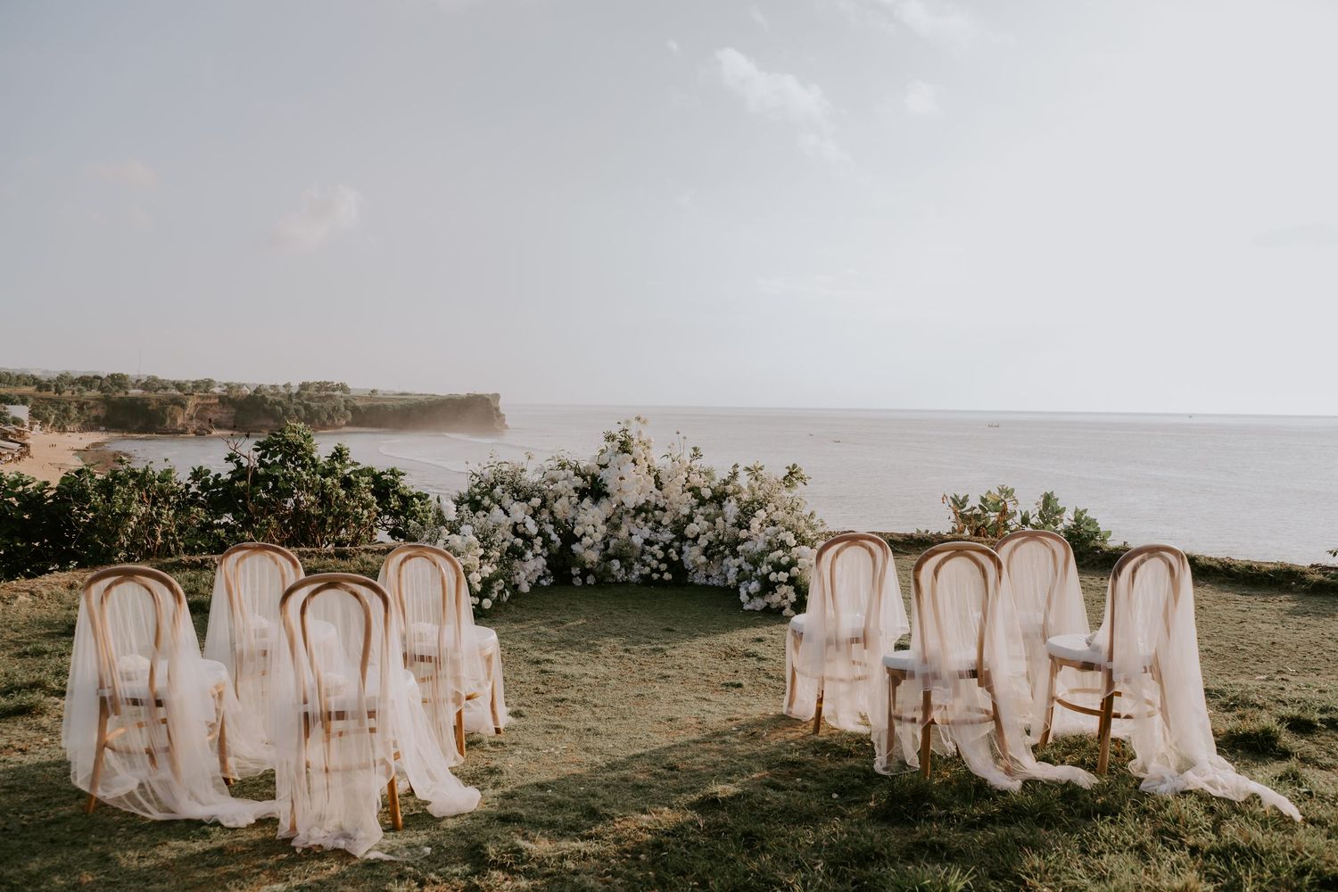 Elegant outdoor wedding ceremony setup with white floral arch overlooking ocean cliffs.