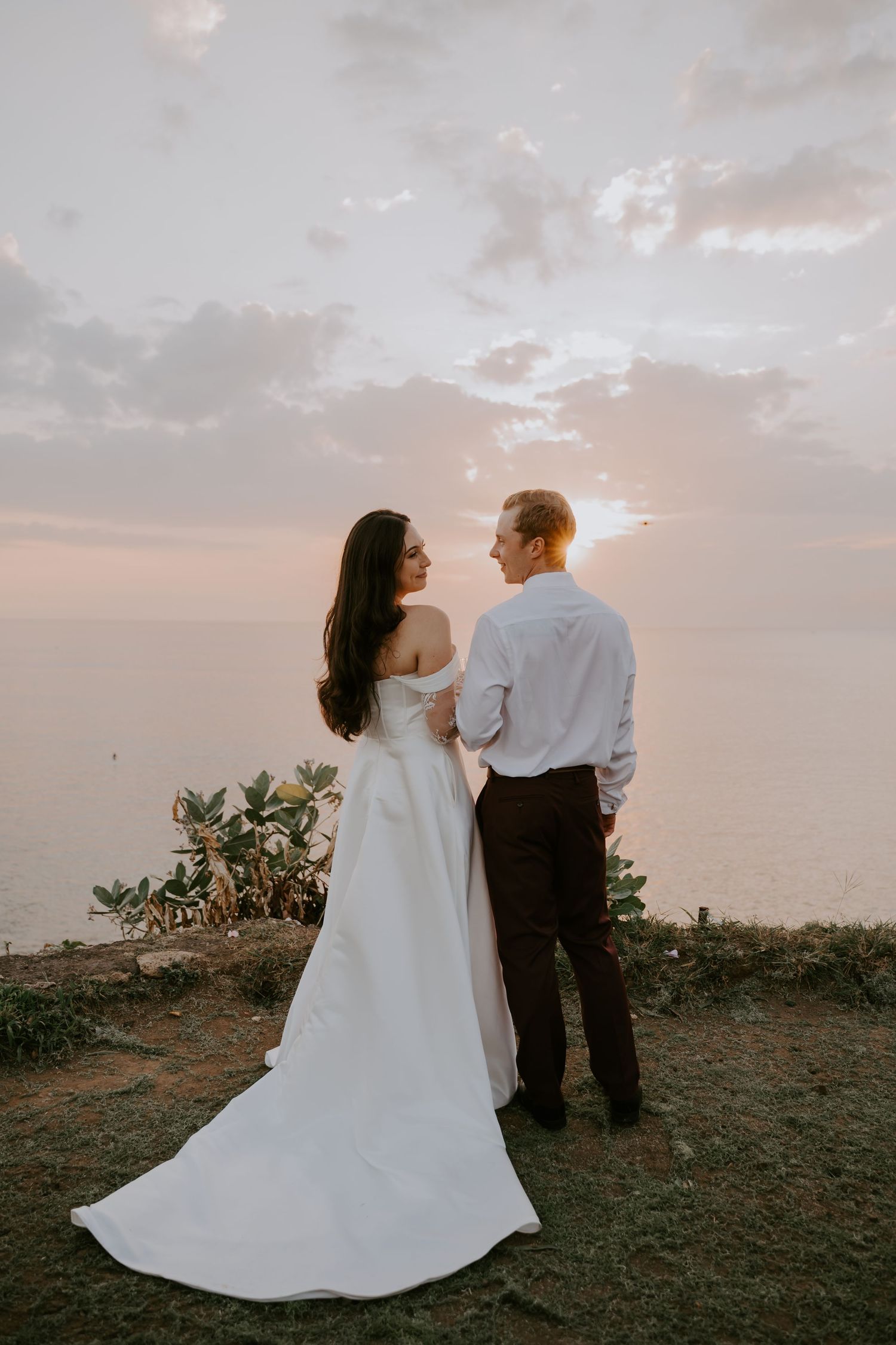 Bride in white off-shoulder gown and groom in white shirt stand together overlooking ocean at sunset.