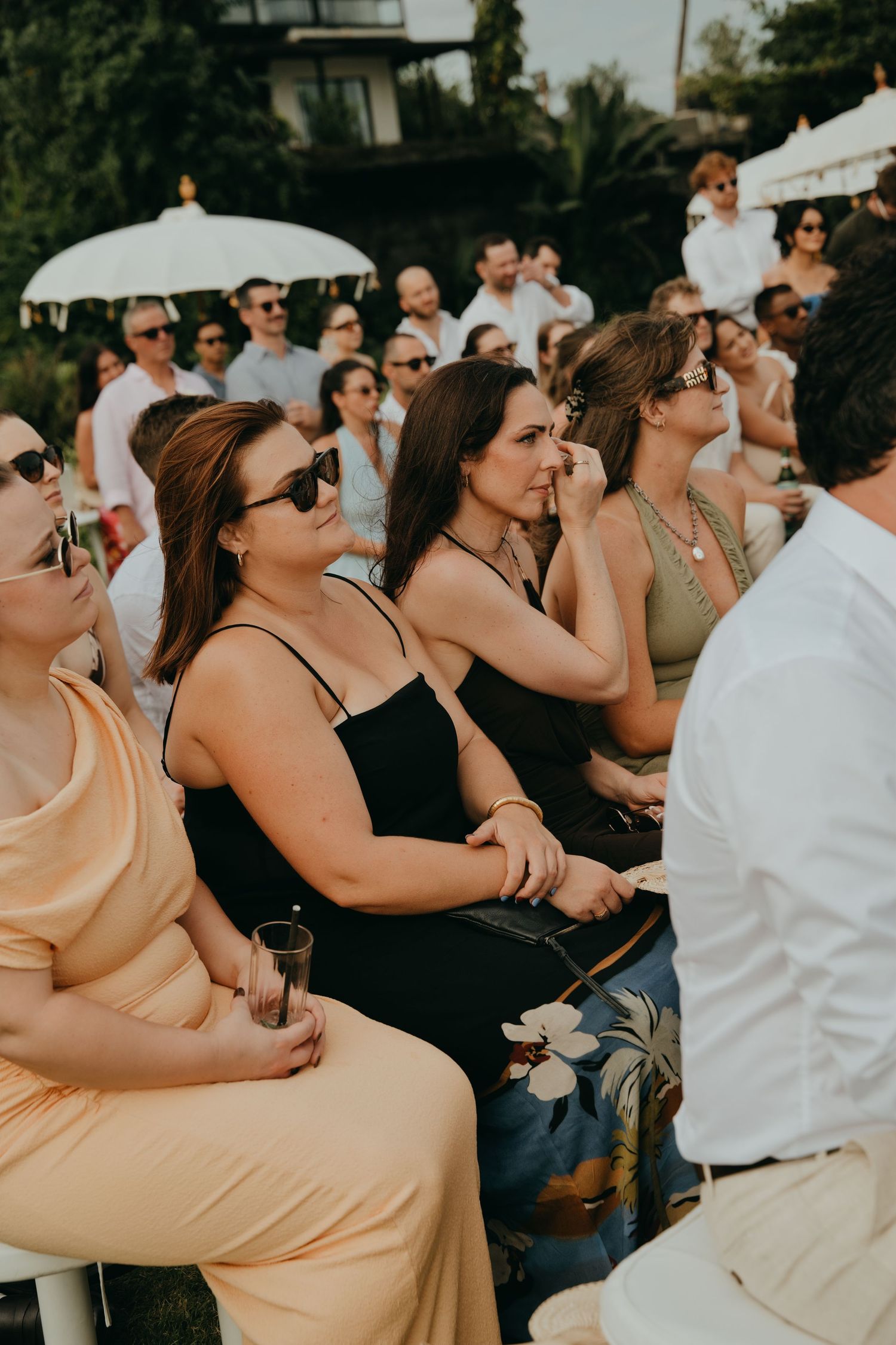 Wedding guests seated outdoors in elegant attire watching a ceremony on a sunny day.