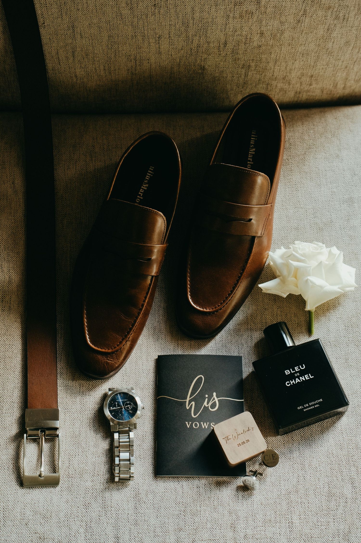 Groom wedding flat lay featuring brown penny loafers, Bleu de Chanel cologne, watch, belt, and vow book.
