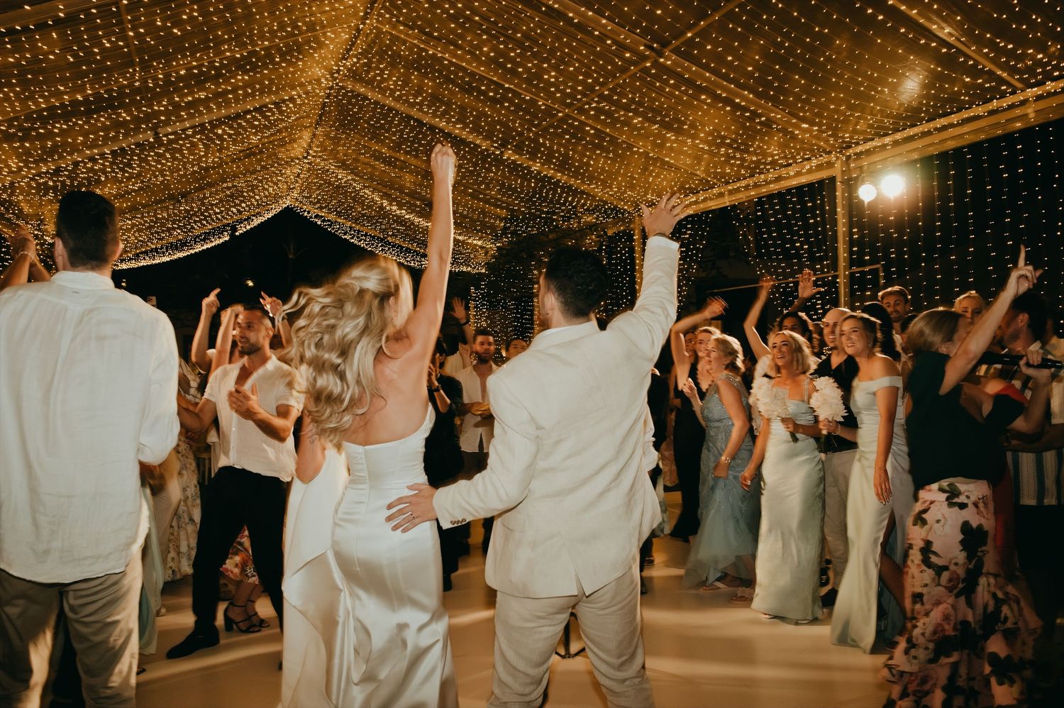 Bride and groom celebrate with hands raised on dance floor under twinkling fairy lights.