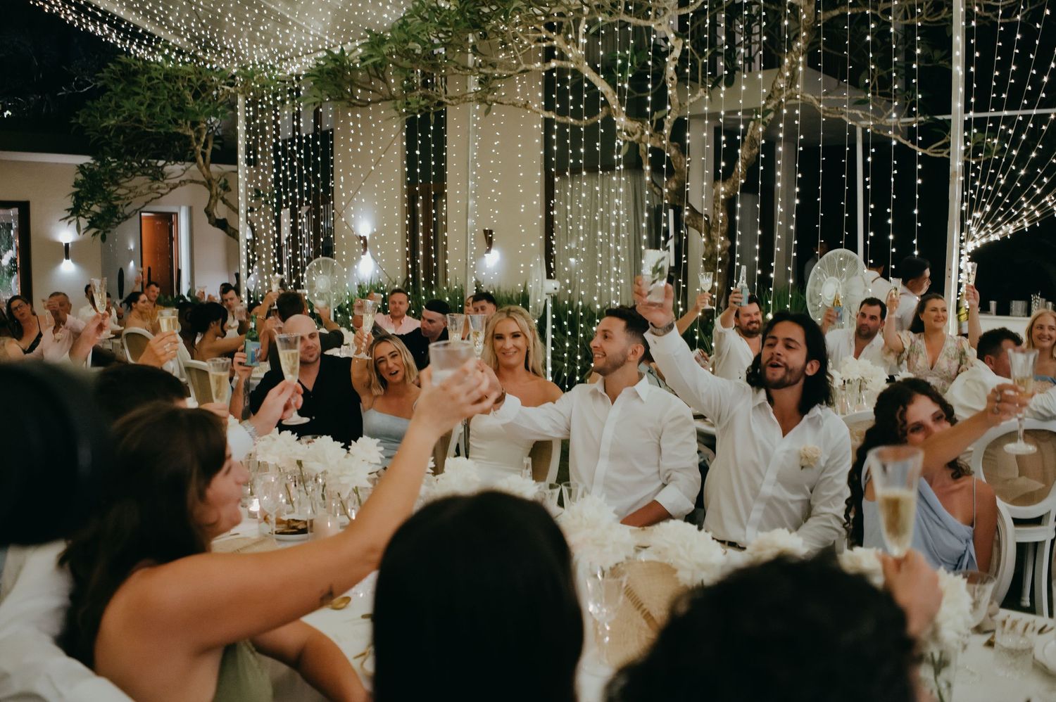 Wedding guests raise glasses in a champagne toast under a canopy of twinkling fairy lights.