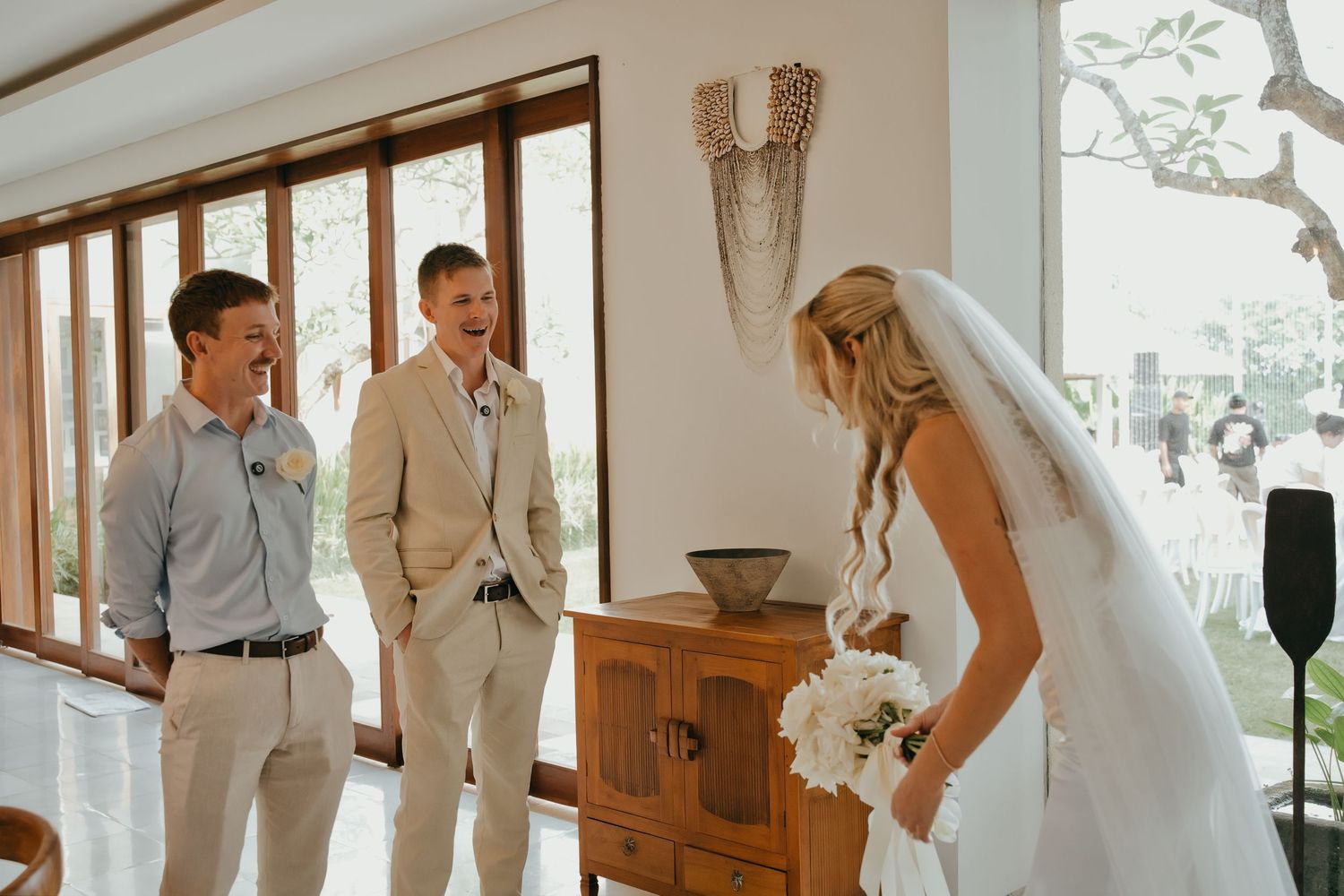 Bride in wedding dress and veil meeting smiling groomsmen during a first look moment indoors.