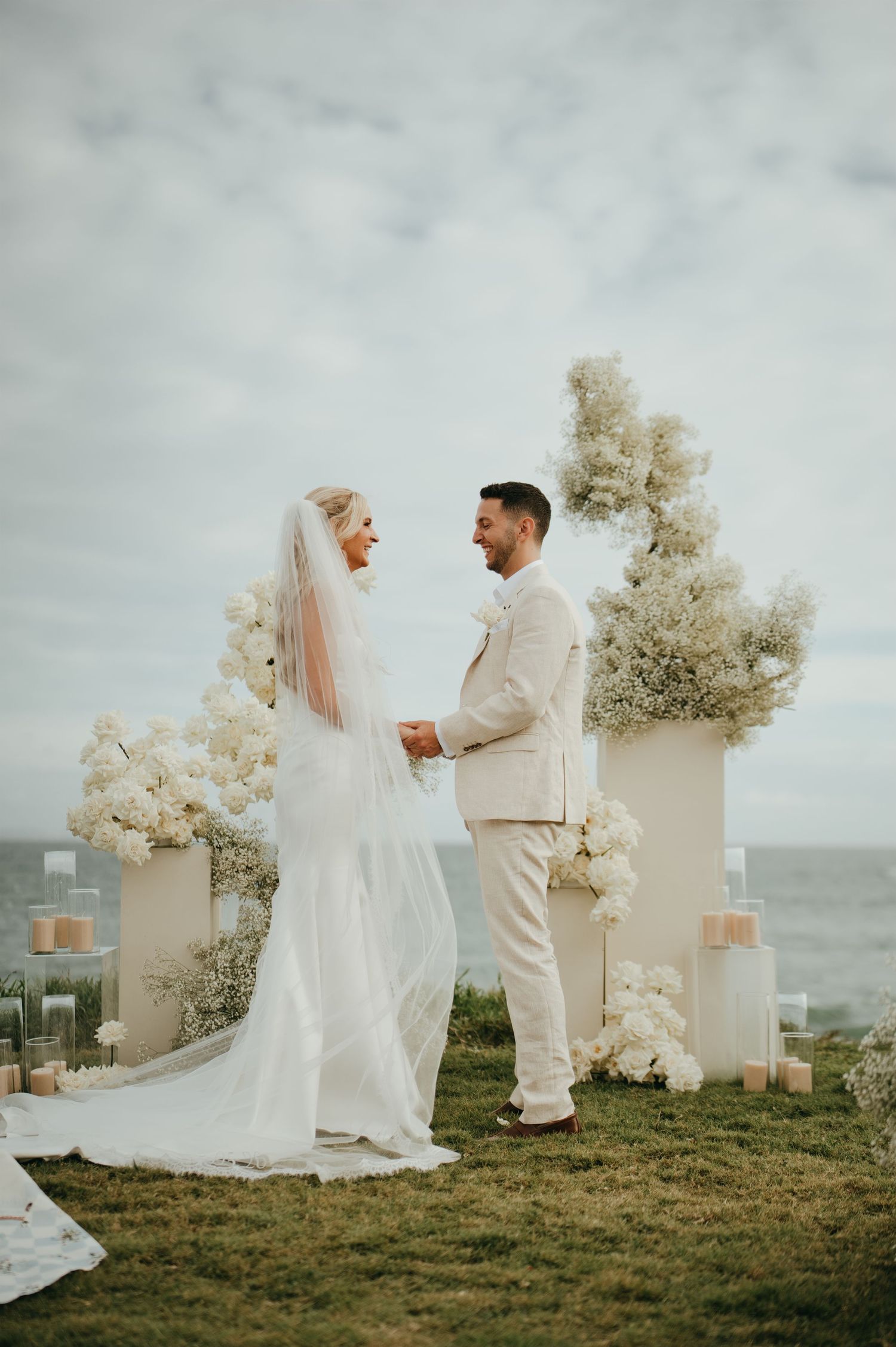 Bride and groom exchanging vows at clifftop ceremony with white floral arch and ocean backdrop.
