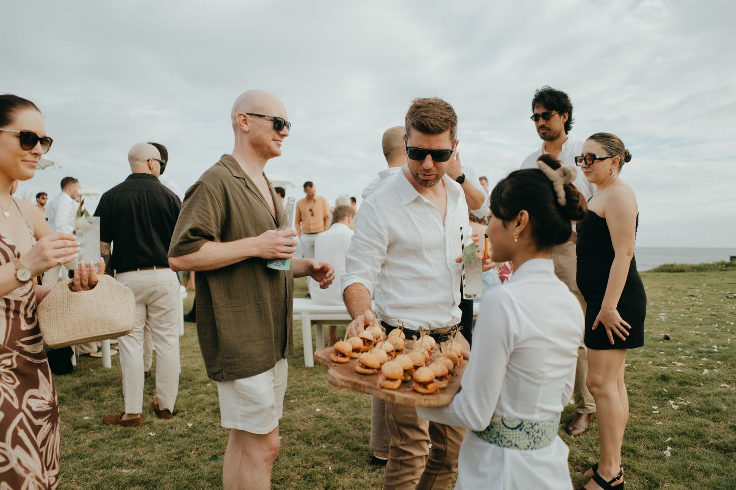 Guests enjoy passed appetizers at an outdoor coastal wedding reception on a grassy cliff.