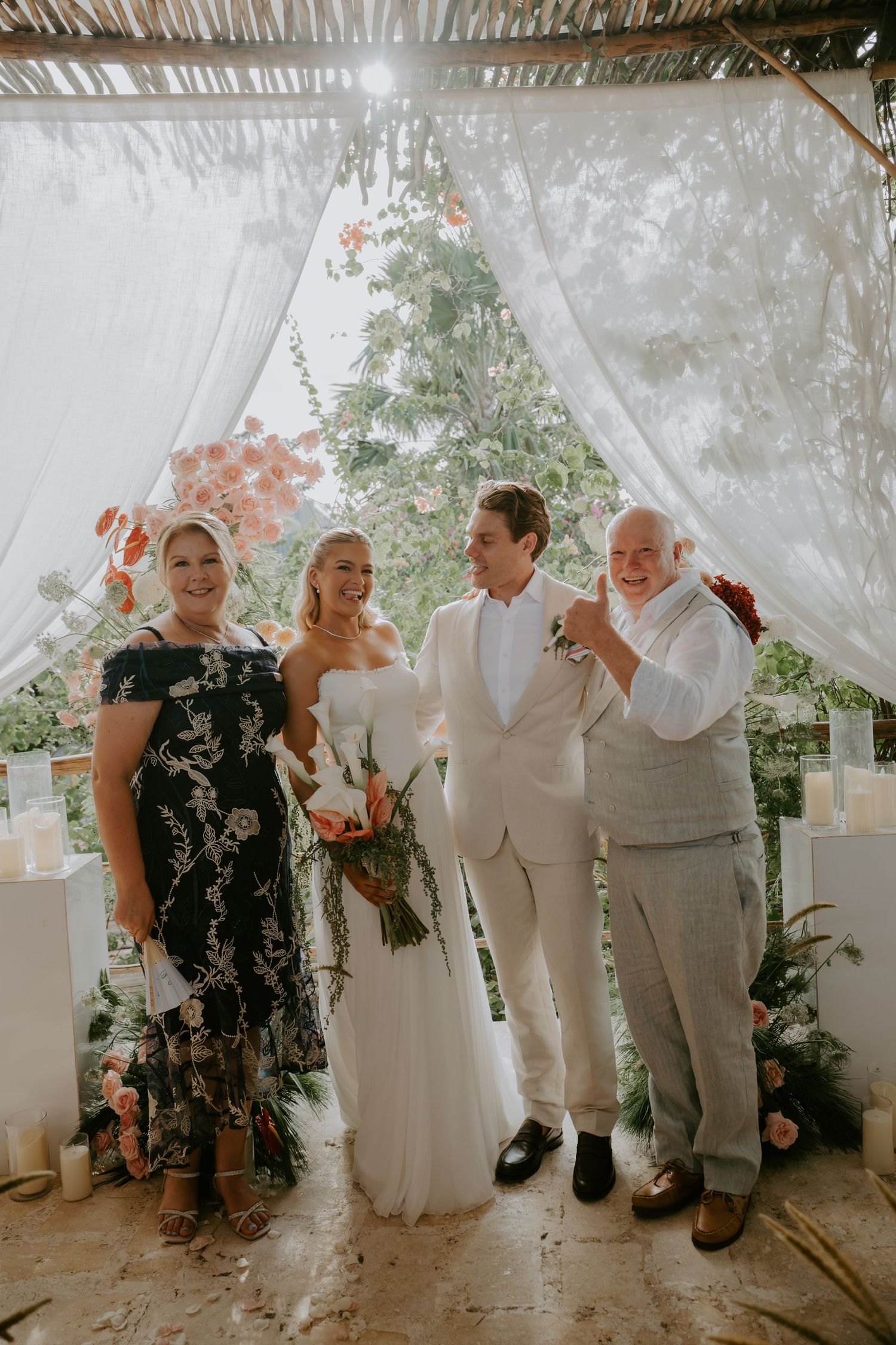 Newlywed couple poses with family members beneath draped altar with candles and tropical floral arch.