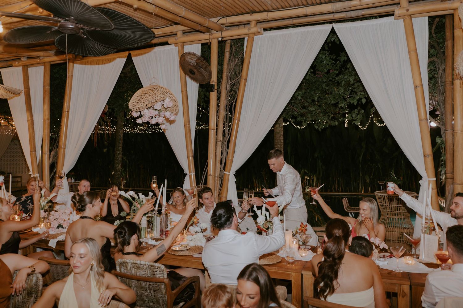 Wedding guests raising glasses in a toast at a tropical bamboo-decorated outdoor reception venue at night.