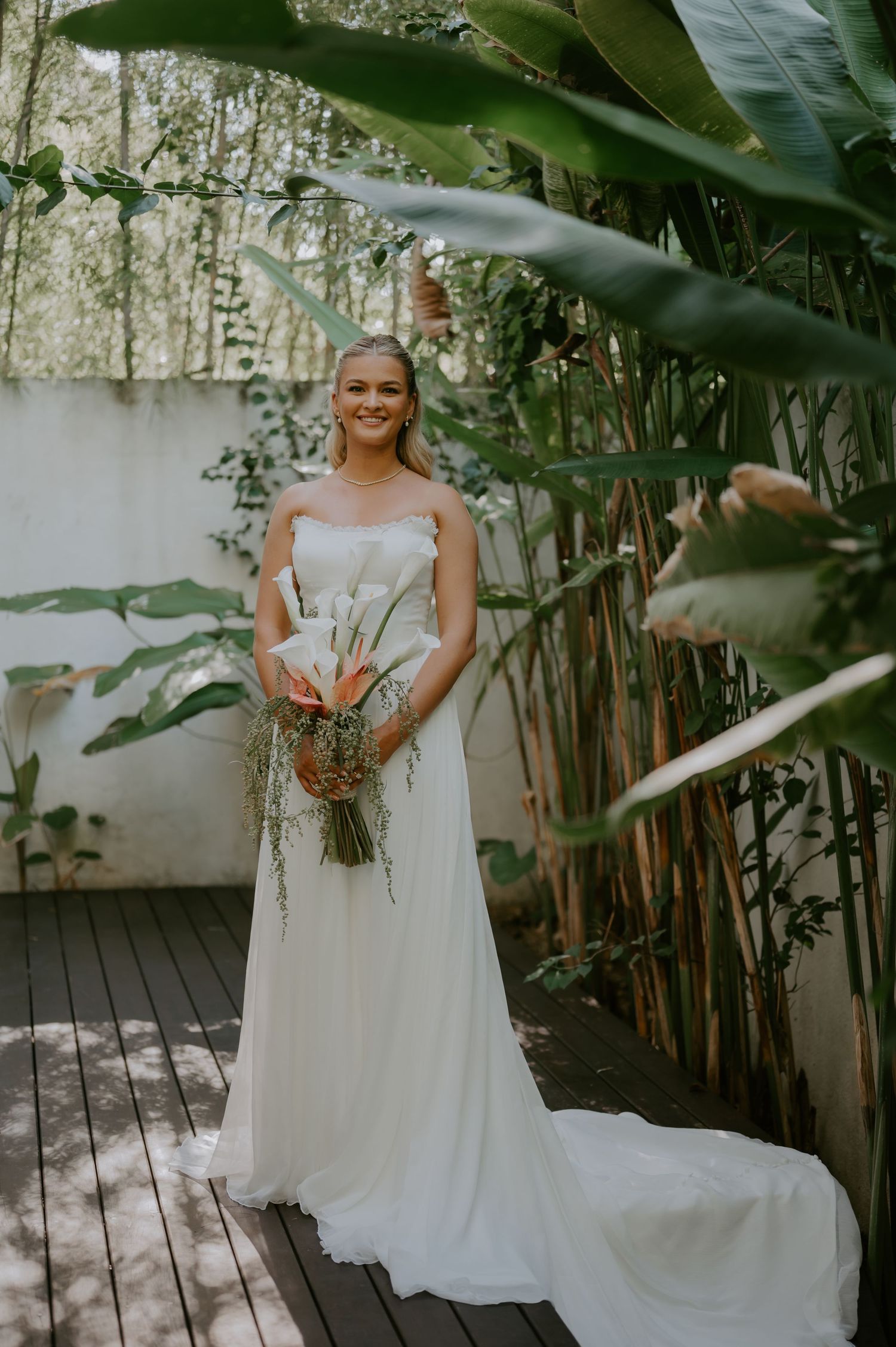Smiling bride in strapless white gown holding tropical bouquet surrounded by lush bamboo and banana leaf greenery.