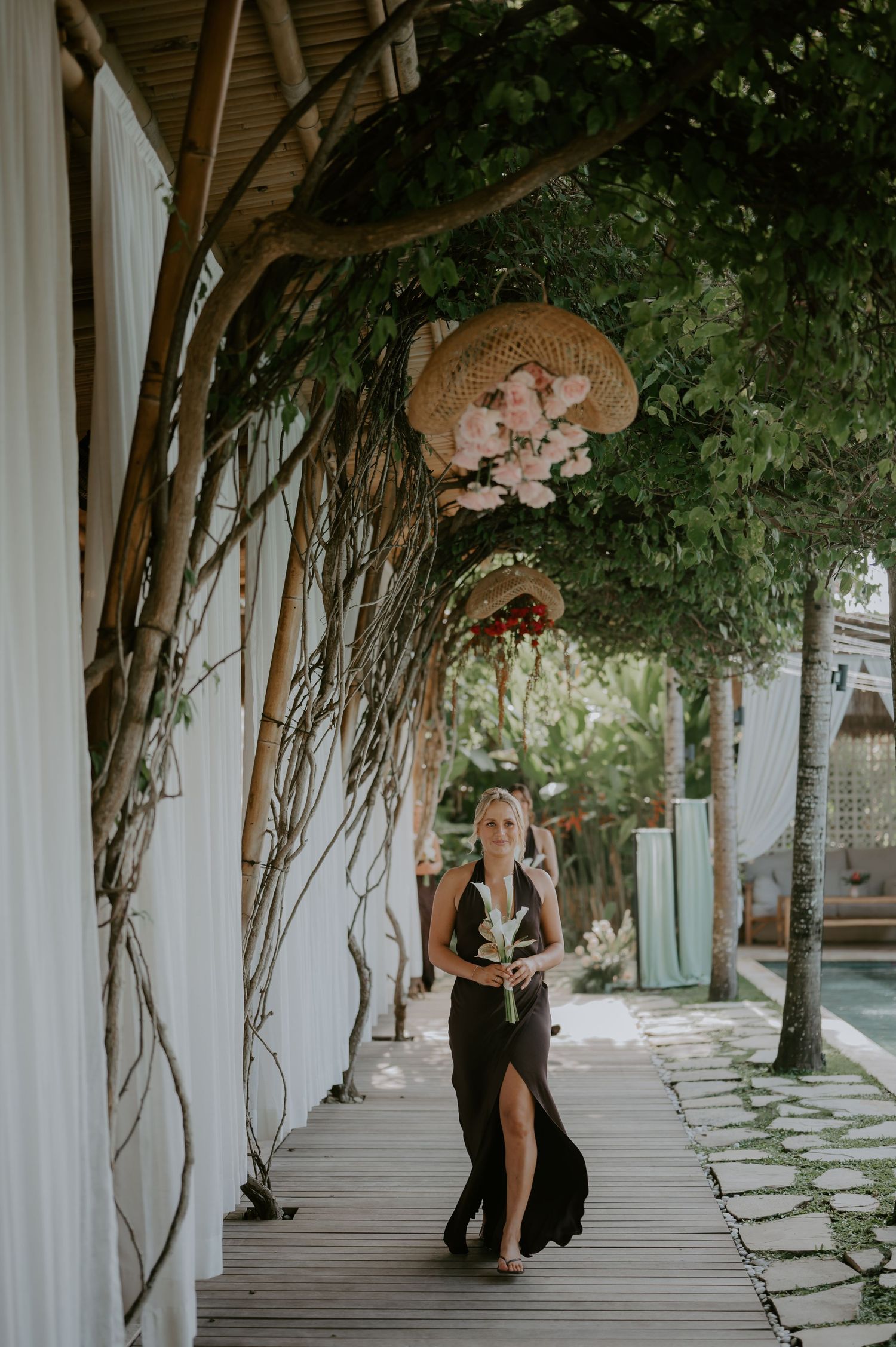 Elegant woman in black maxi dress walks under floral straw hat arch on a wooden pathway surrounded by greenery.