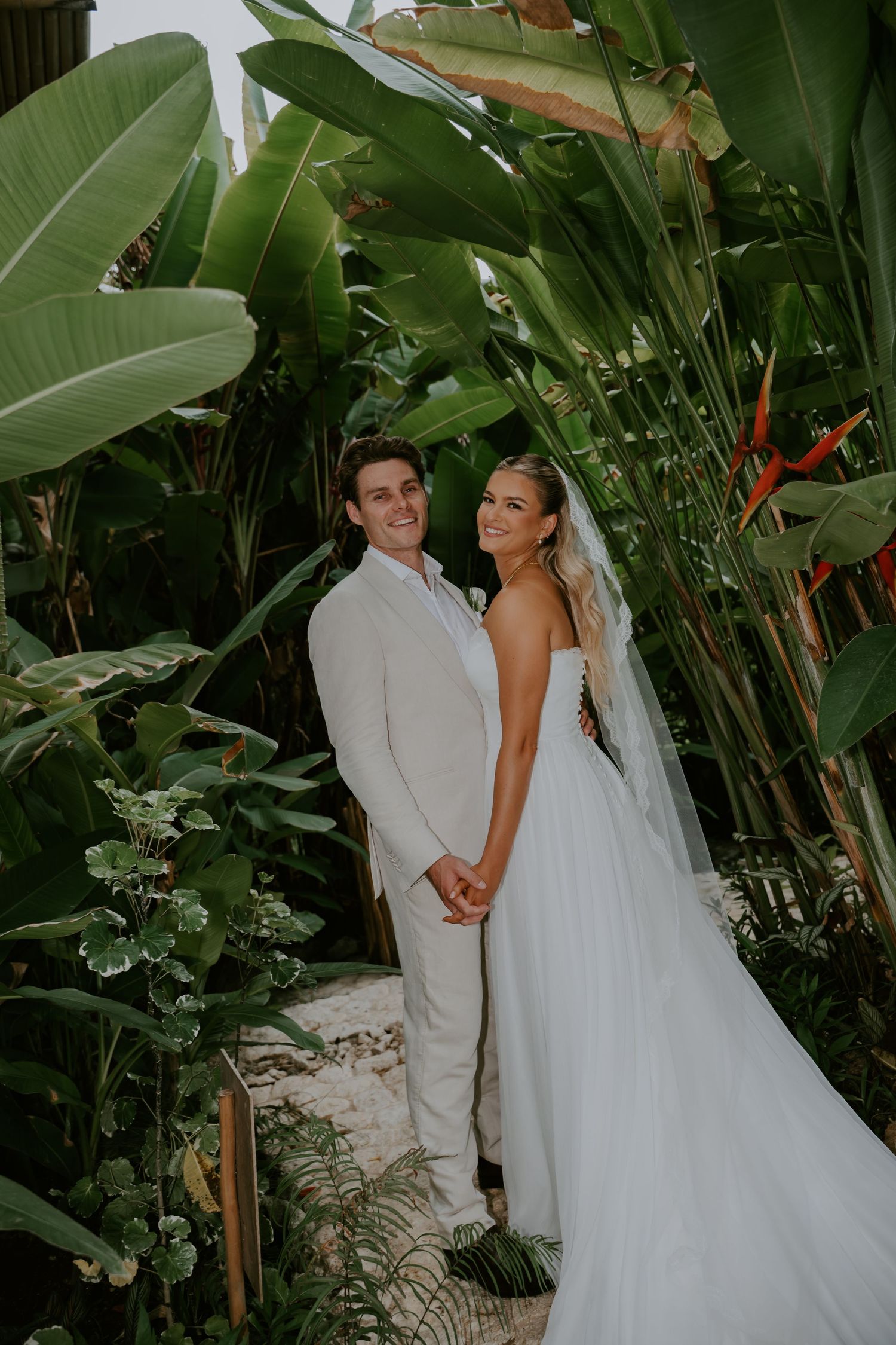 Smiling newlyweds hold hands surrounded by lush tropical banana leaf plants after outdoor wedding.