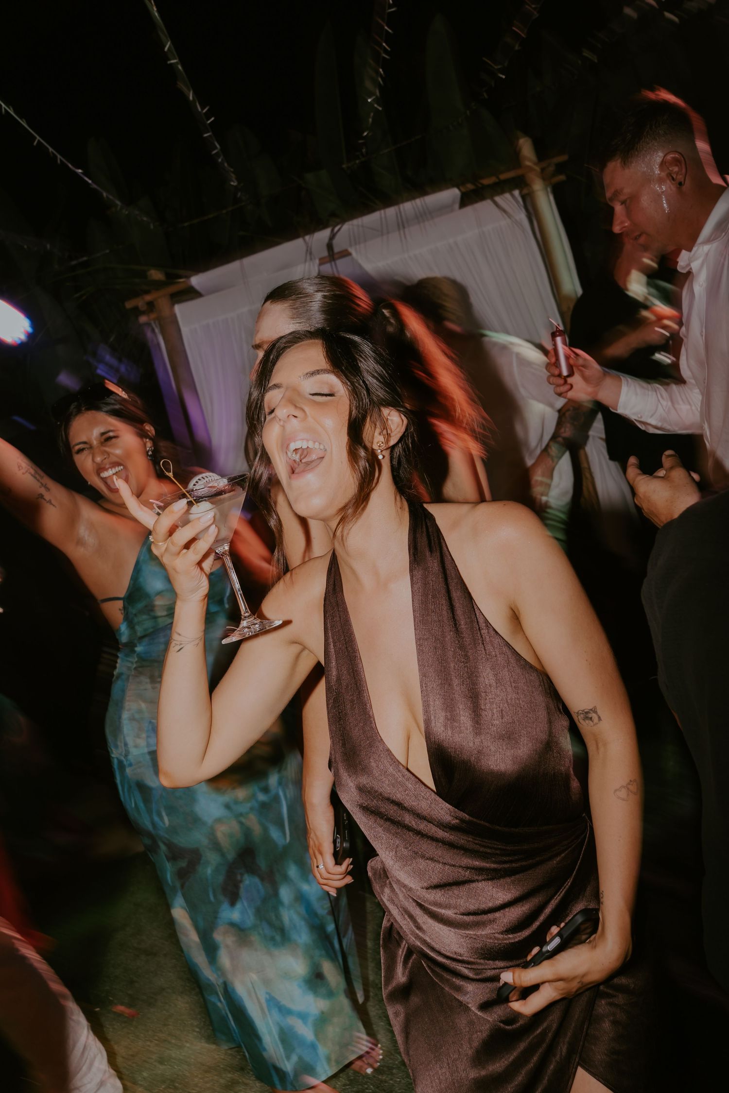 Wedding guest in elegant halter dress dancing with a cocktail at outdoor evening reception.