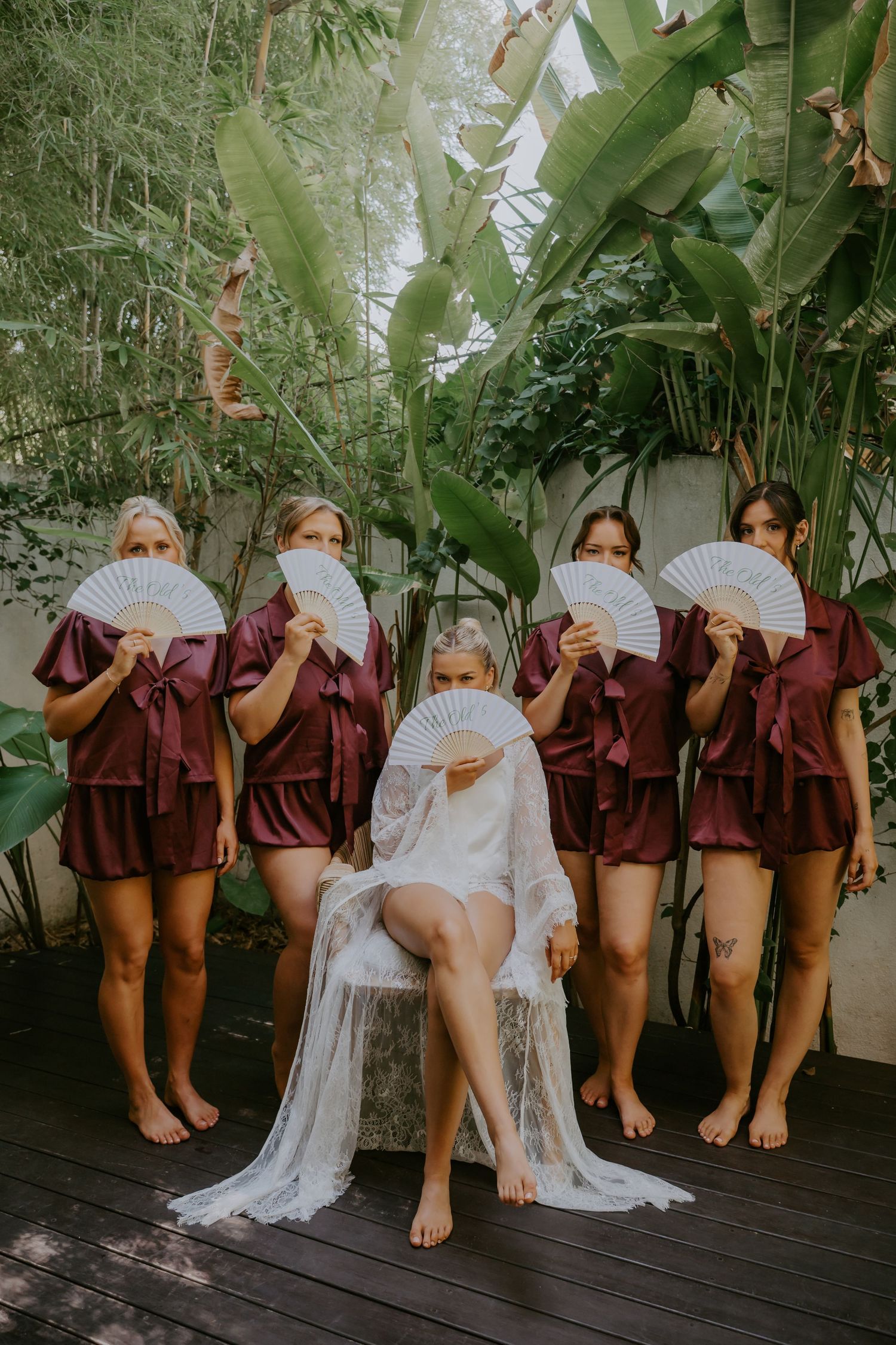 Bride in white lace robe sits with bridesmaids in burgundy robes holding white fans outdoors.