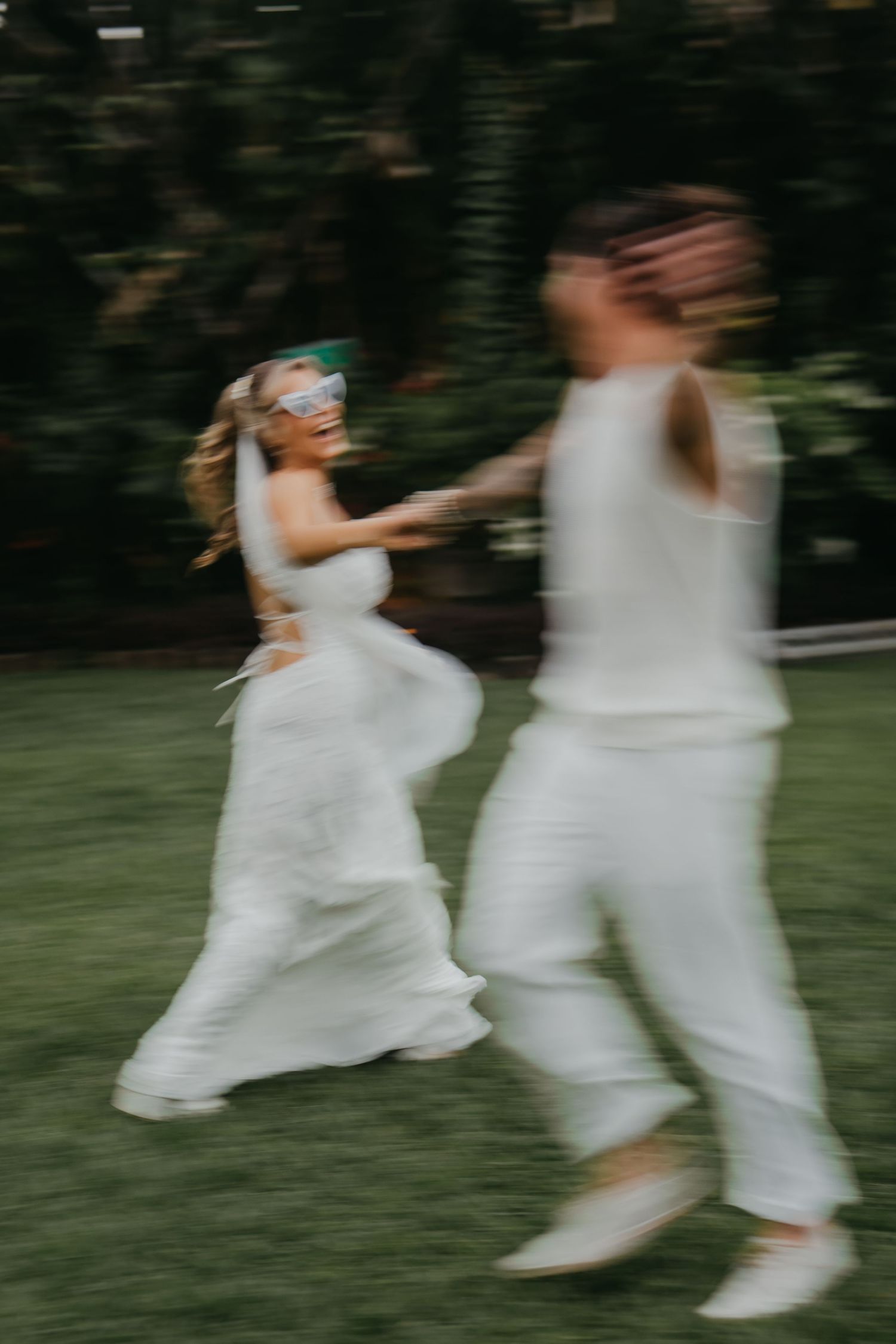 Two brides in white holding hands and running joyfully across a green lawn on their wedding day.