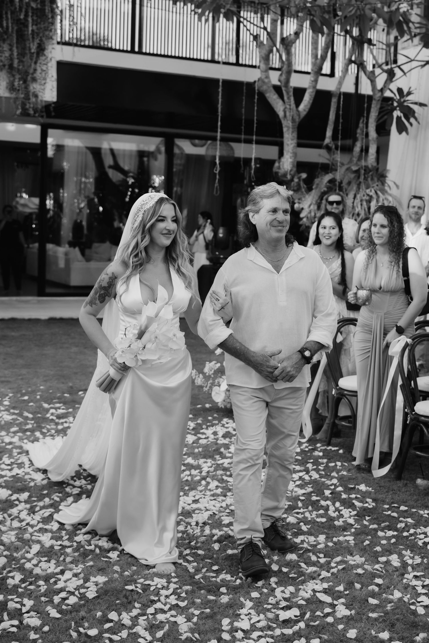 Black and white photo of bride walking down aisle with father past flower petal covered ground.